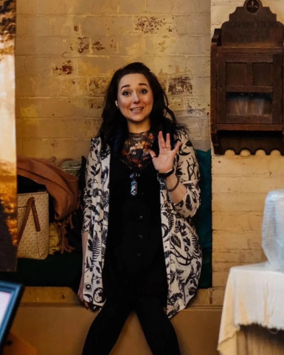 A woman with long dark hair smiling and waving, sitting on a bench against a rustic wooden wall in a warmly lit indoor setting.