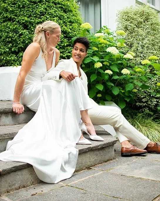 Two women in white dresses sit on outdoor steps, smiling and having a conversation, with green shrubs and flowering plants in the background.