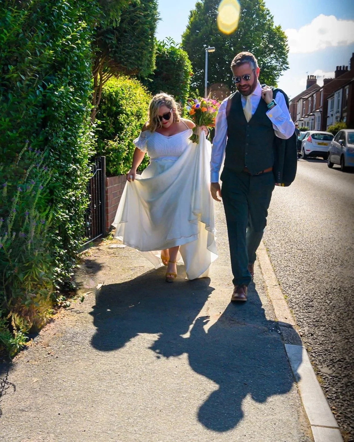 A woman in a white wedding dress holding a bouquet and a man in a suit walking on a sidewalk on a sunny day. They are both wearing sunglasses, and there are cars parked along the street. Green bushes and trees are visible on the left side of the sidewalk.
