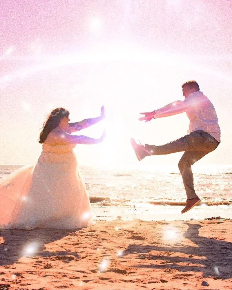 A couple on the beach at sunset, with the man jumping mid-air and the woman facing him, both with outstretched arms, in a playful pose.