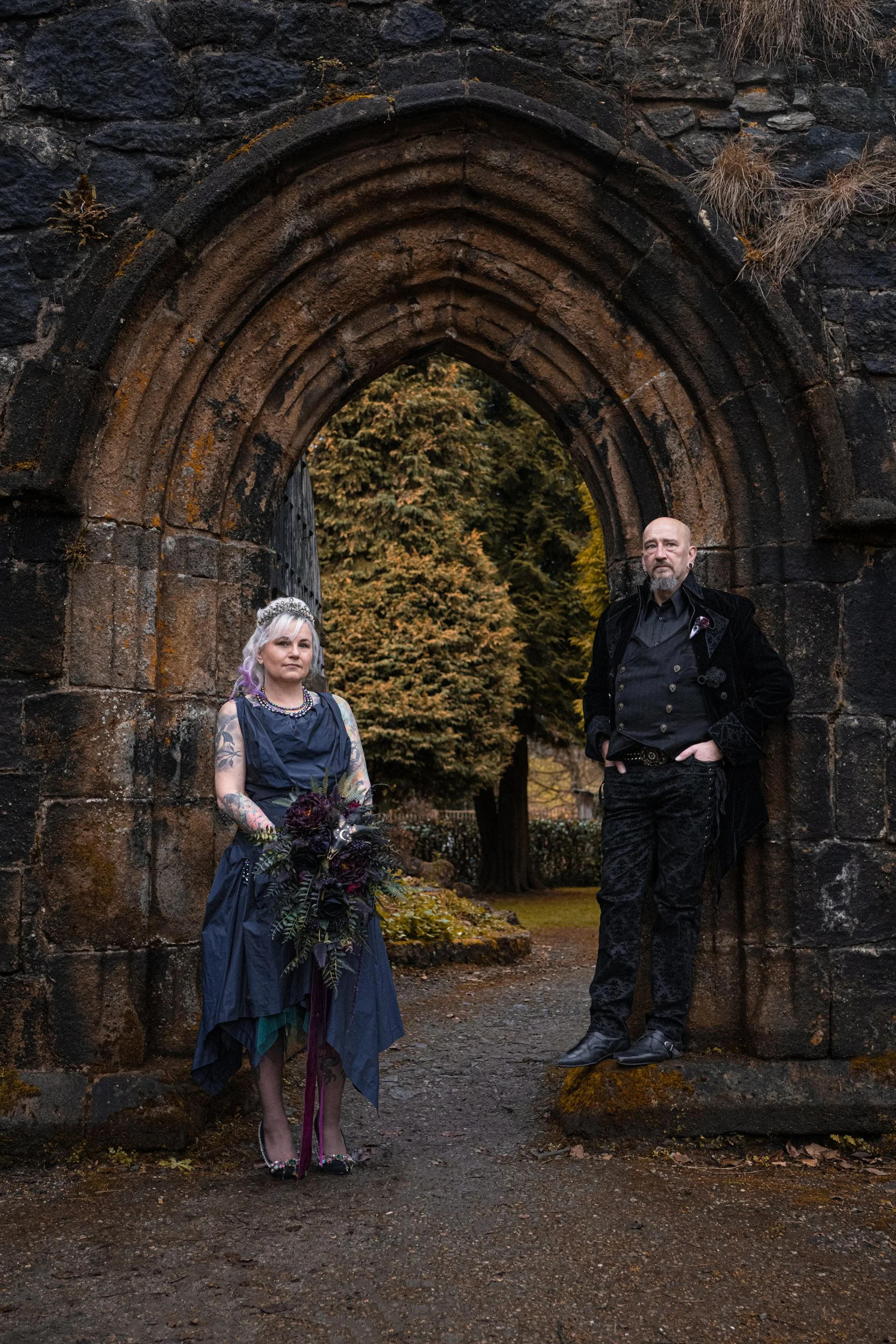 A gothic couple standing in a gothic archway within some castle ruins. The wife stands to the left and the husband to the right, both in black wedding attire staring down the camera. post-wedding shoot