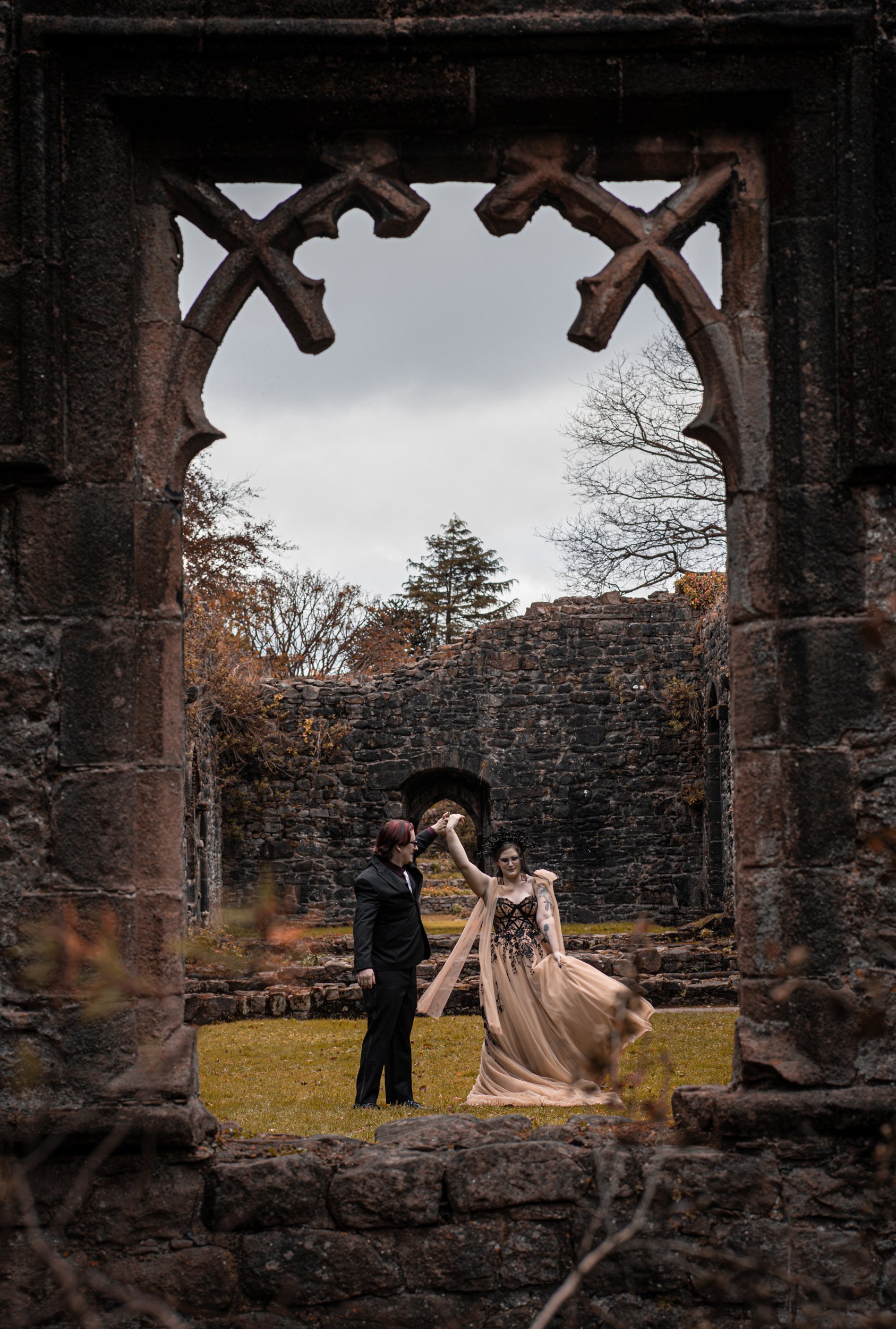 A Husband and Wife dancing in castle ruins. The image for their Post-Wedding shoot being captured through a window. The husband if wearing a black suit and the Wife is wearing a Legend Bridal Dress, gold and black in colour.