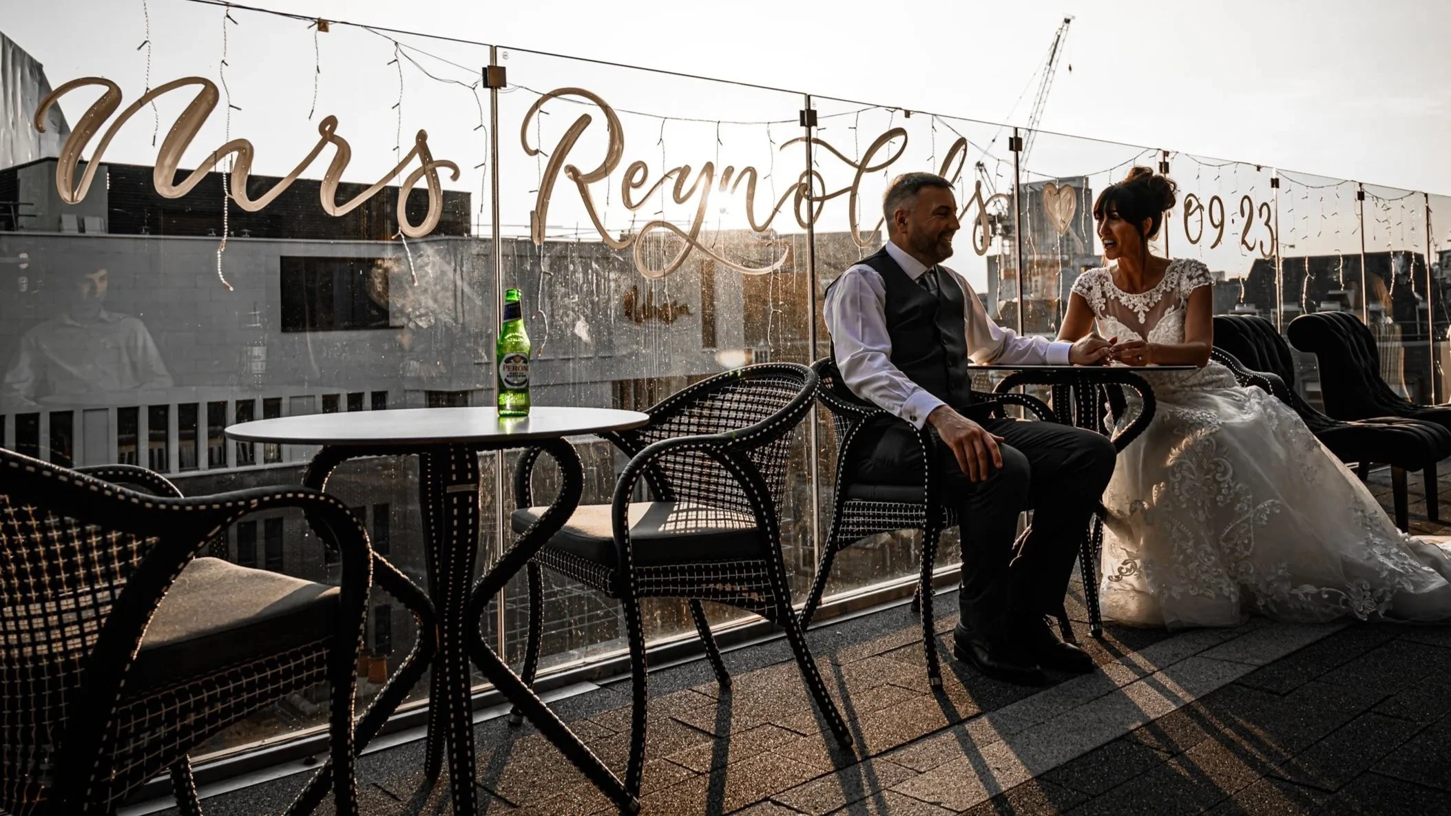 A bride and groom sitting together on a rooftop terrace, smiling and holding hands, with wedding decorations and sunlight in the background.
