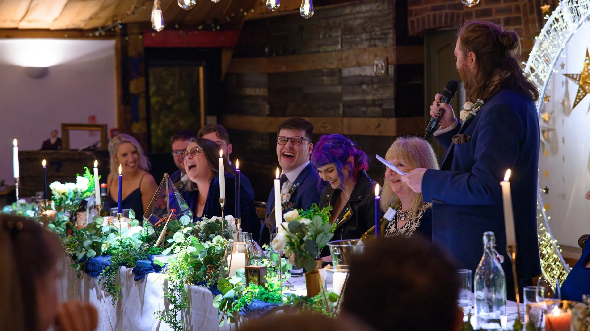 wedding day speeches. Everyone at the top table is hysterically laughing. The bride and groom look embraced. Bride has pink and purple hair with a custom leather jacket. Groom has blue suit.