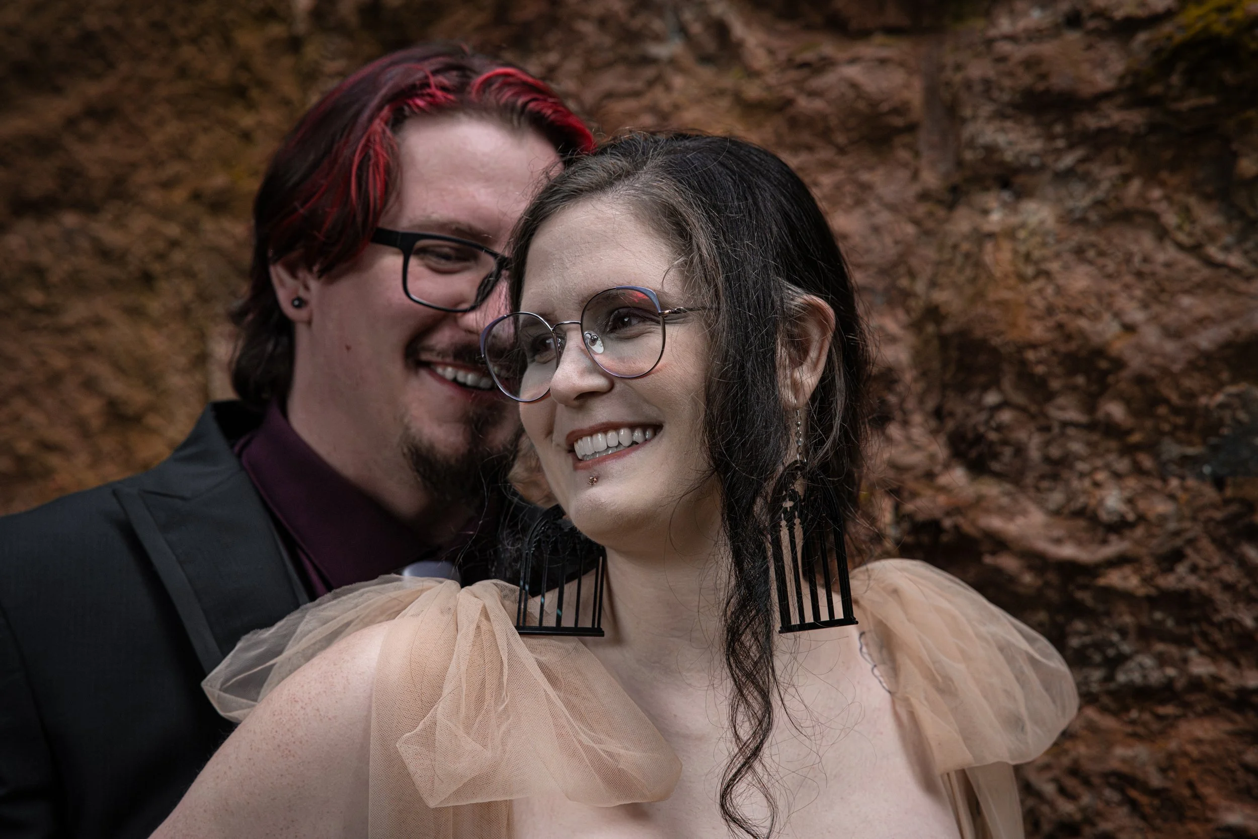 A photo of a couple smiling, the husband behind the wife. Both with big smiles on their faces. The wife is wearing Cathedral window earrings.