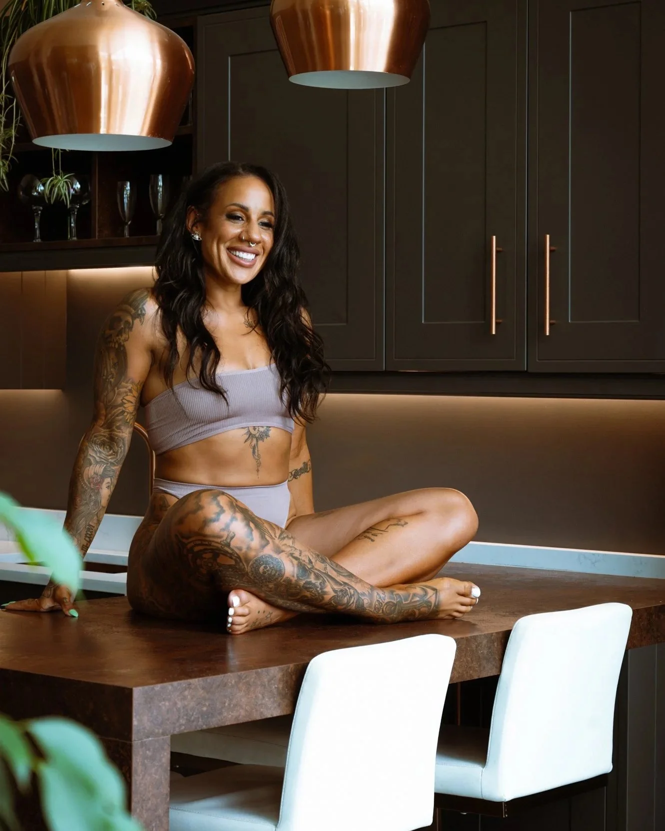A woman with tattoos sitting on a kitchen table in a modern kitchen, smiling. She is wearing a light purple sports bra and matching shorts, with aromatic lighting and dark cabinets in the background.