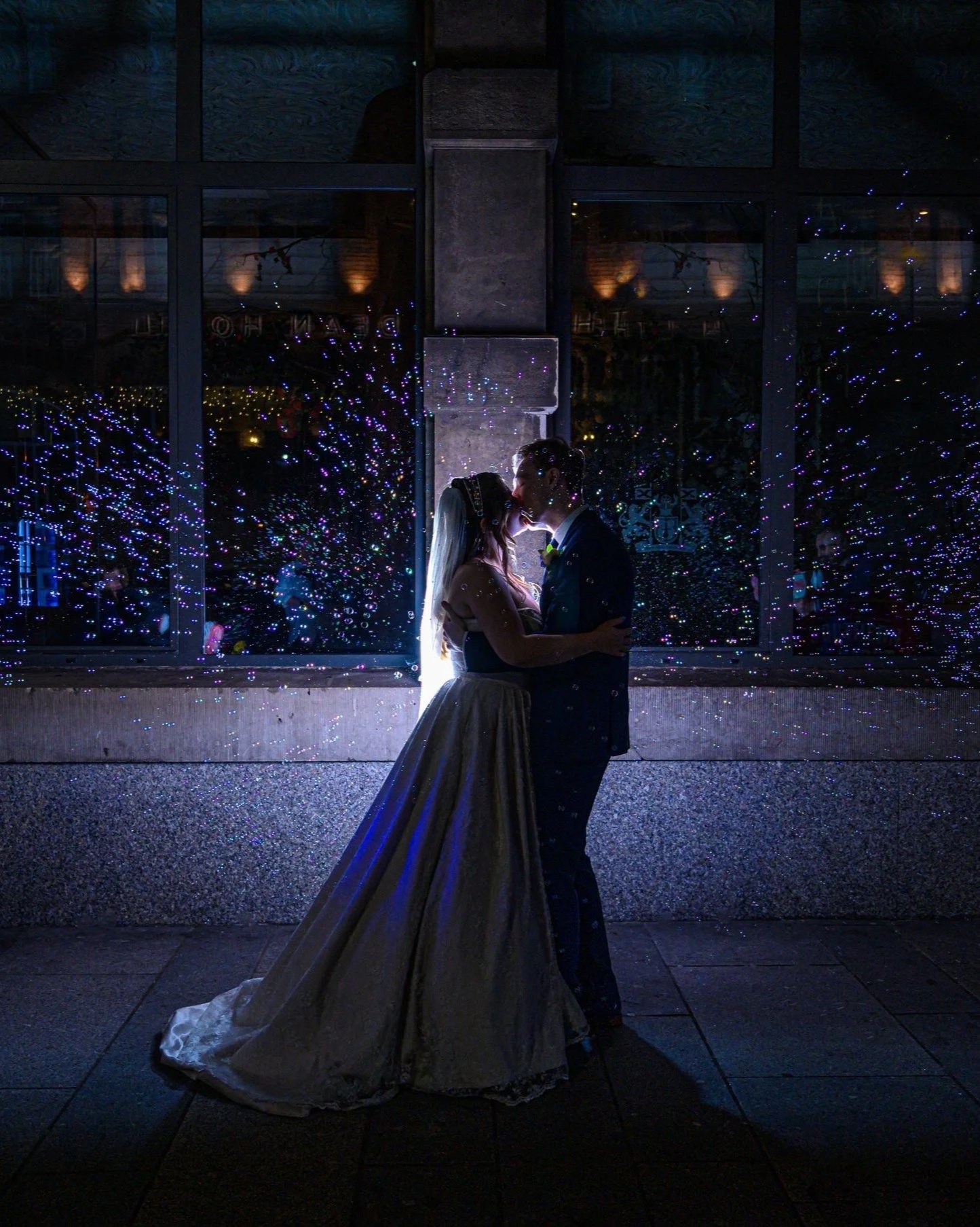 A bride and groom share a romantic kiss at night, silhouetted against a large window decorated with purple and blue fairy lights, with reflections of warm indoor lighting visible behind the glass.