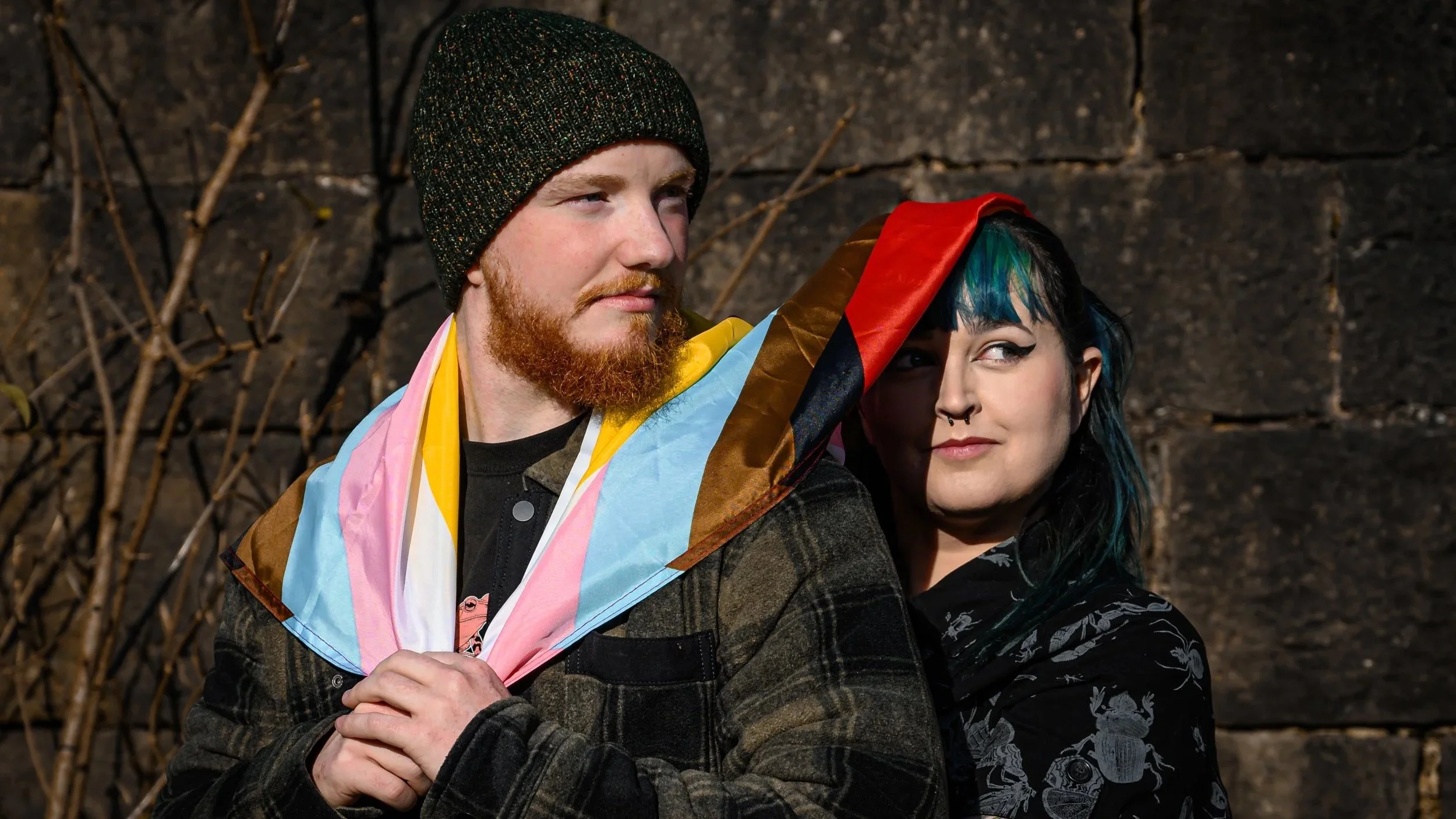 LGBTQ couple on their engagement shoot being silly and purposefully awkward. Pride flag as a cape and mock serious faces. Transgender man dressed like a skater boy, and bisexual woman with nose perching and blue/green hair.