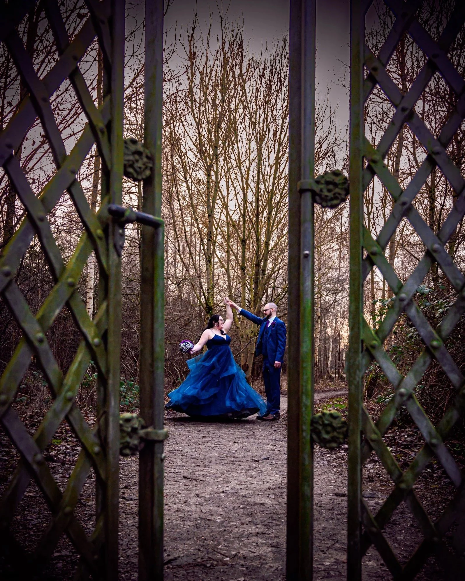 A couple stands holding hands in a wooded outdoor area during sunset, with the woman wearing a long blue gown and holding a bouquet, and the man in a dark suit. The photo is taken through a metal fence.