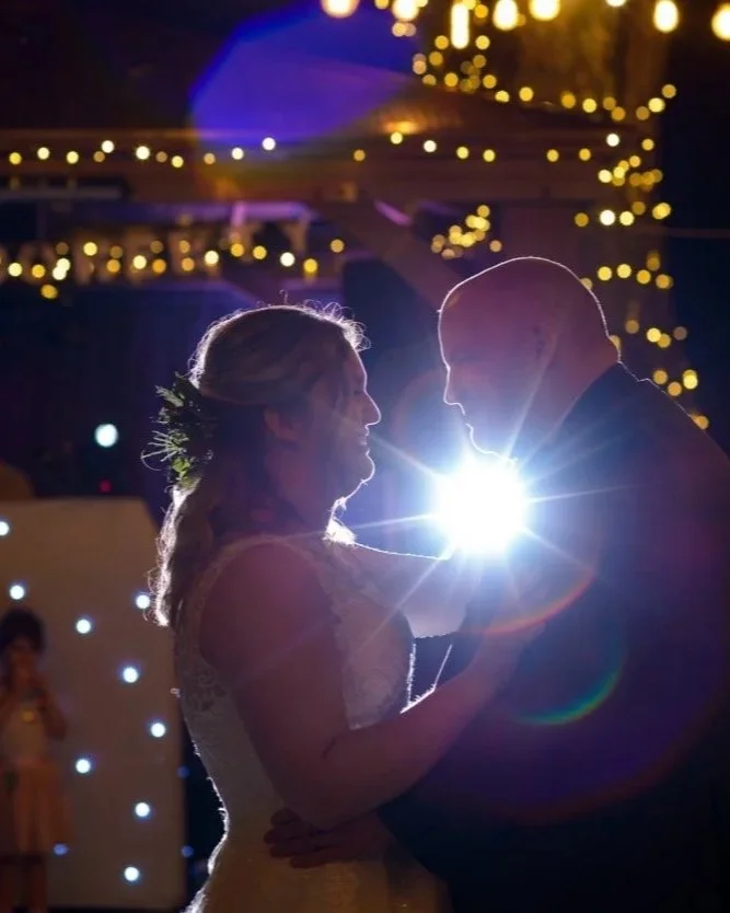 A bride and groom sharing a dance at their wedding with bright stage lights and fairy lights in the background, creating a romantic silhouette.