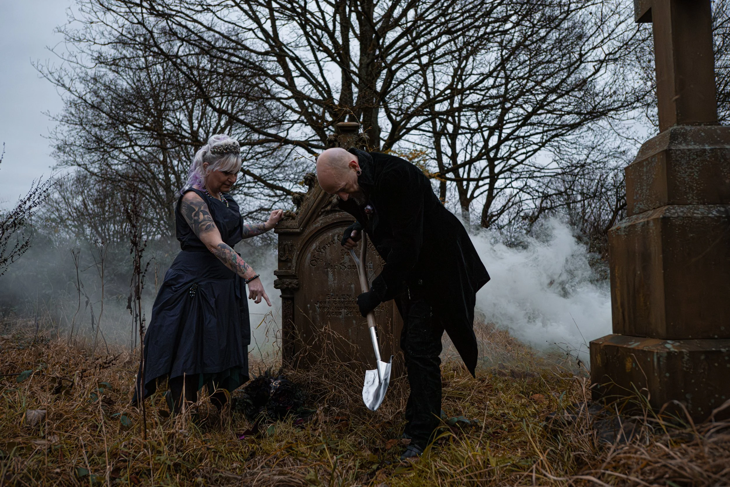 An alternative couple, the wife in a black wedding dress pionting to where her husband, wearing a tailed black velvet suit, should dig. They're in an abandoned cemetery. Fake, all entirely for their post-wedding shoot.