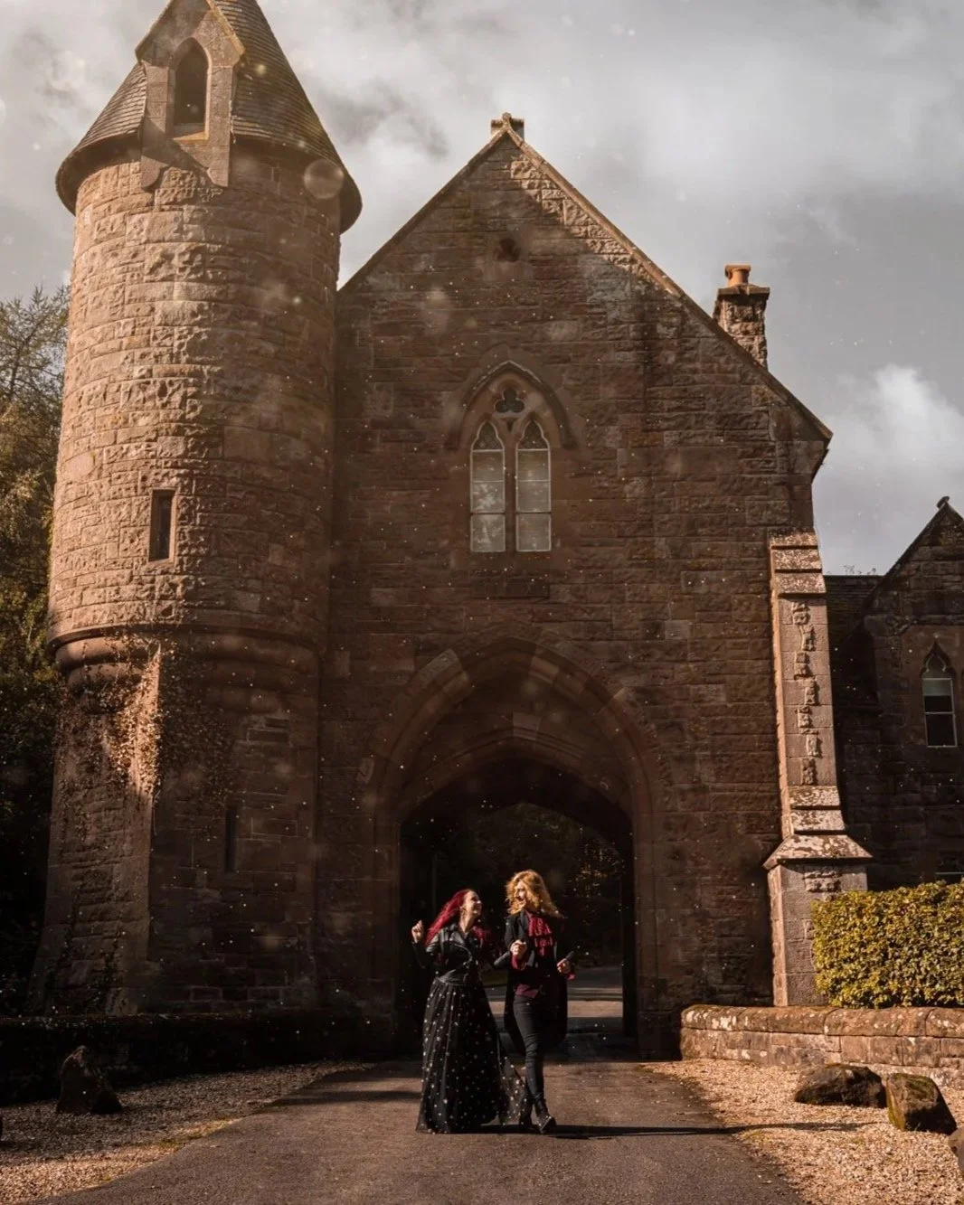 Two women walking and chatting in front of a historic stone castle with a turretted tower, arched entrance, and Gothic windows. The scene is outdoors on a paved pathway during daytime with a cloudy sky.