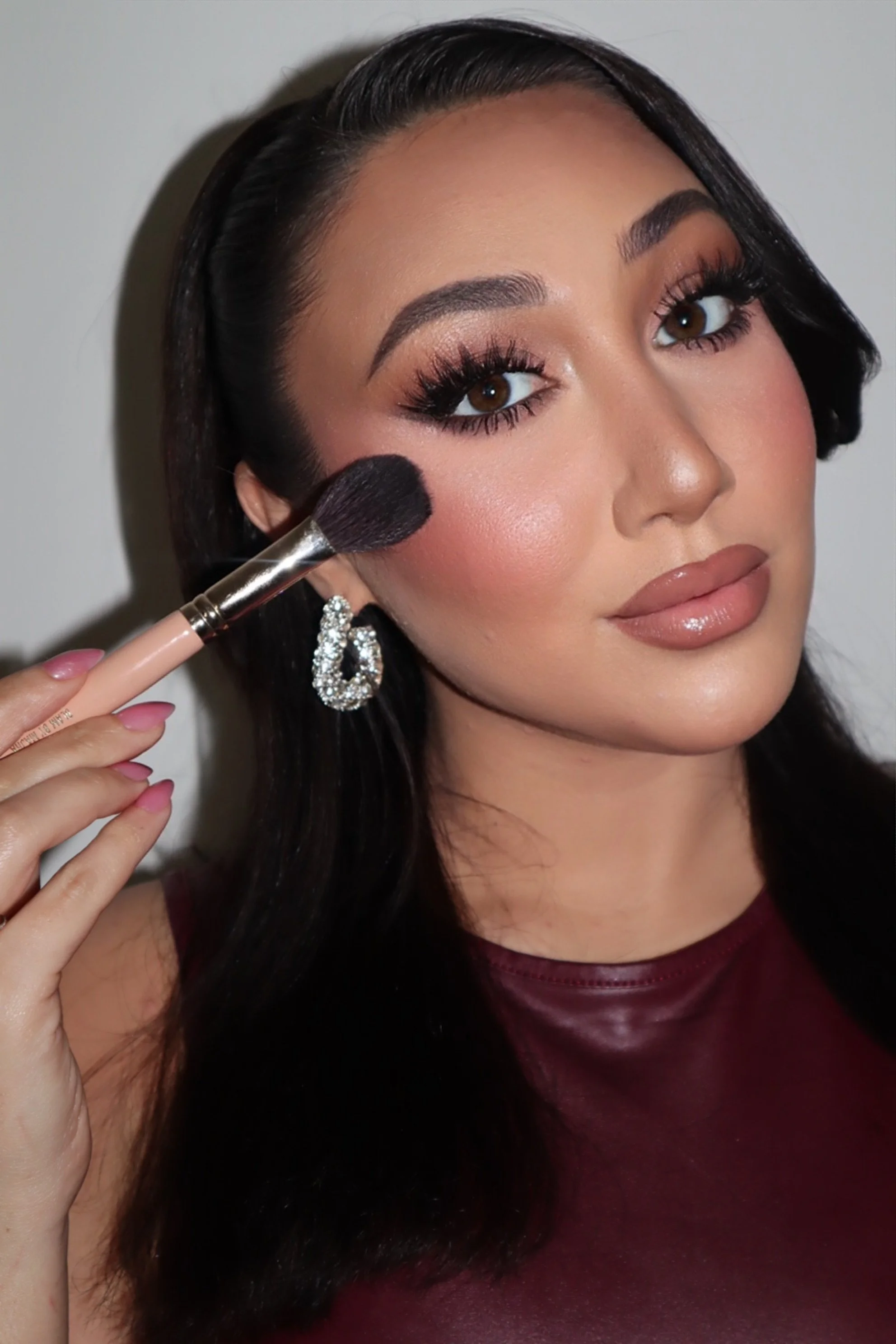 A woman with dark hair and makeup applying blush with a makeup brush.