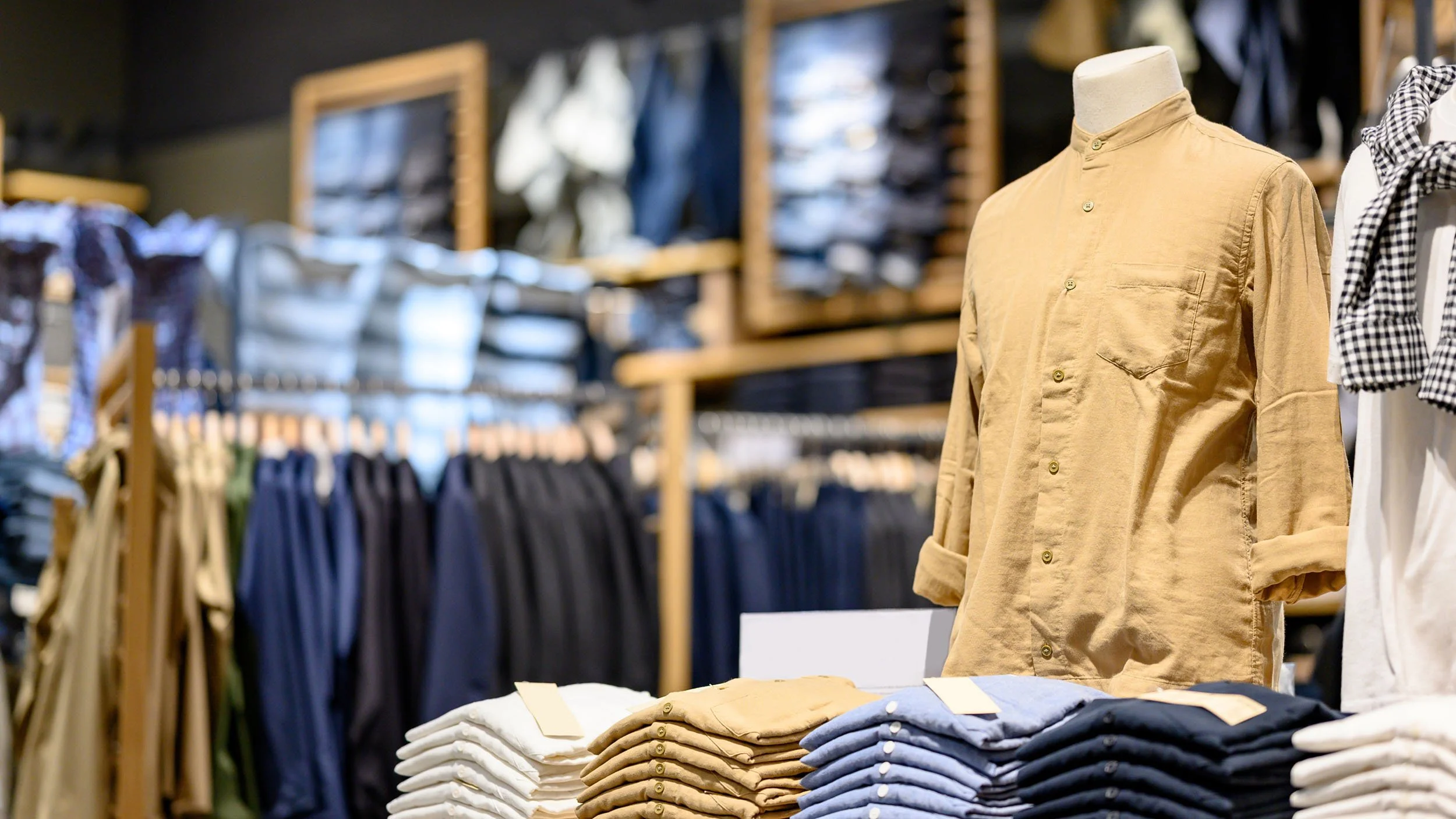 Display of folded khaki, white, and blue pants on a table in a clothing store, with a beige shirt on a mannequin and racks of clothes in the background.