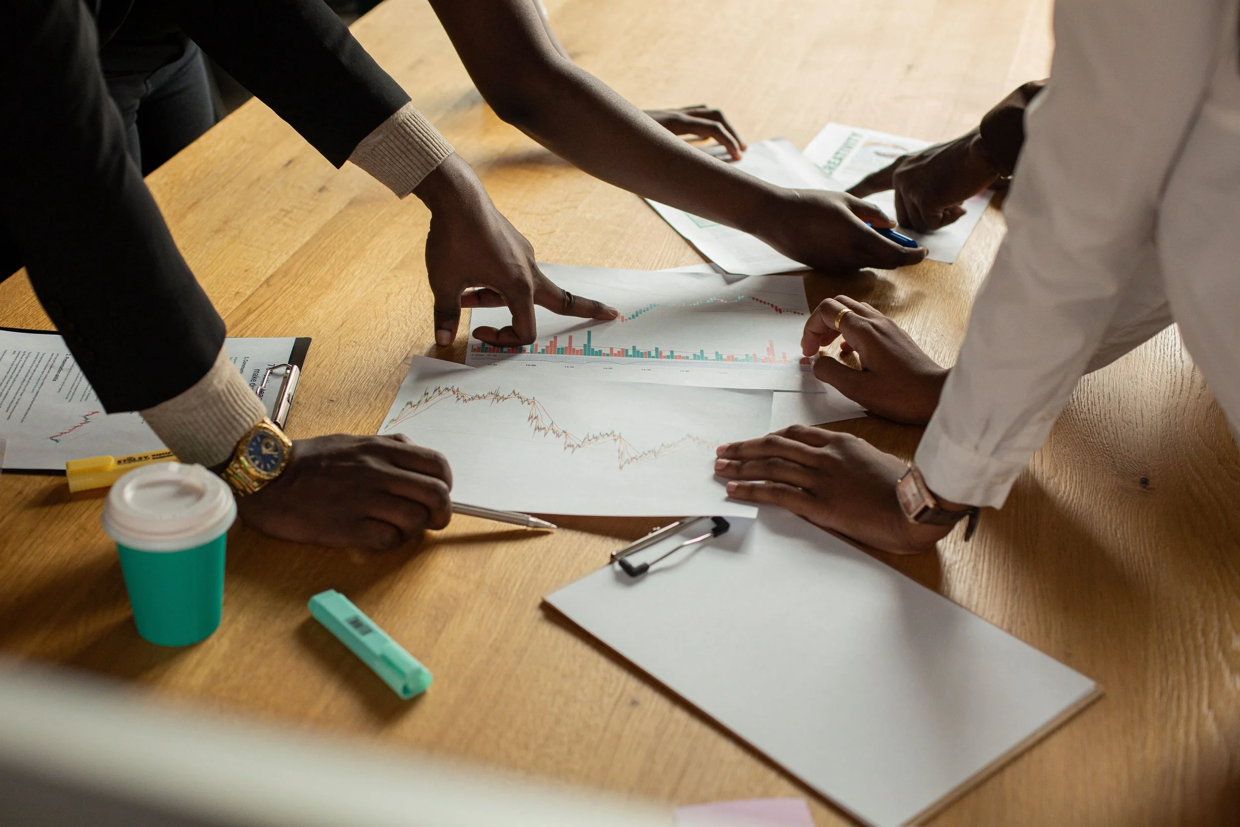 Multiple people are gathered around a wooden table, examining and discussing printed charts and graphs, with pens and a clipboard on the table.