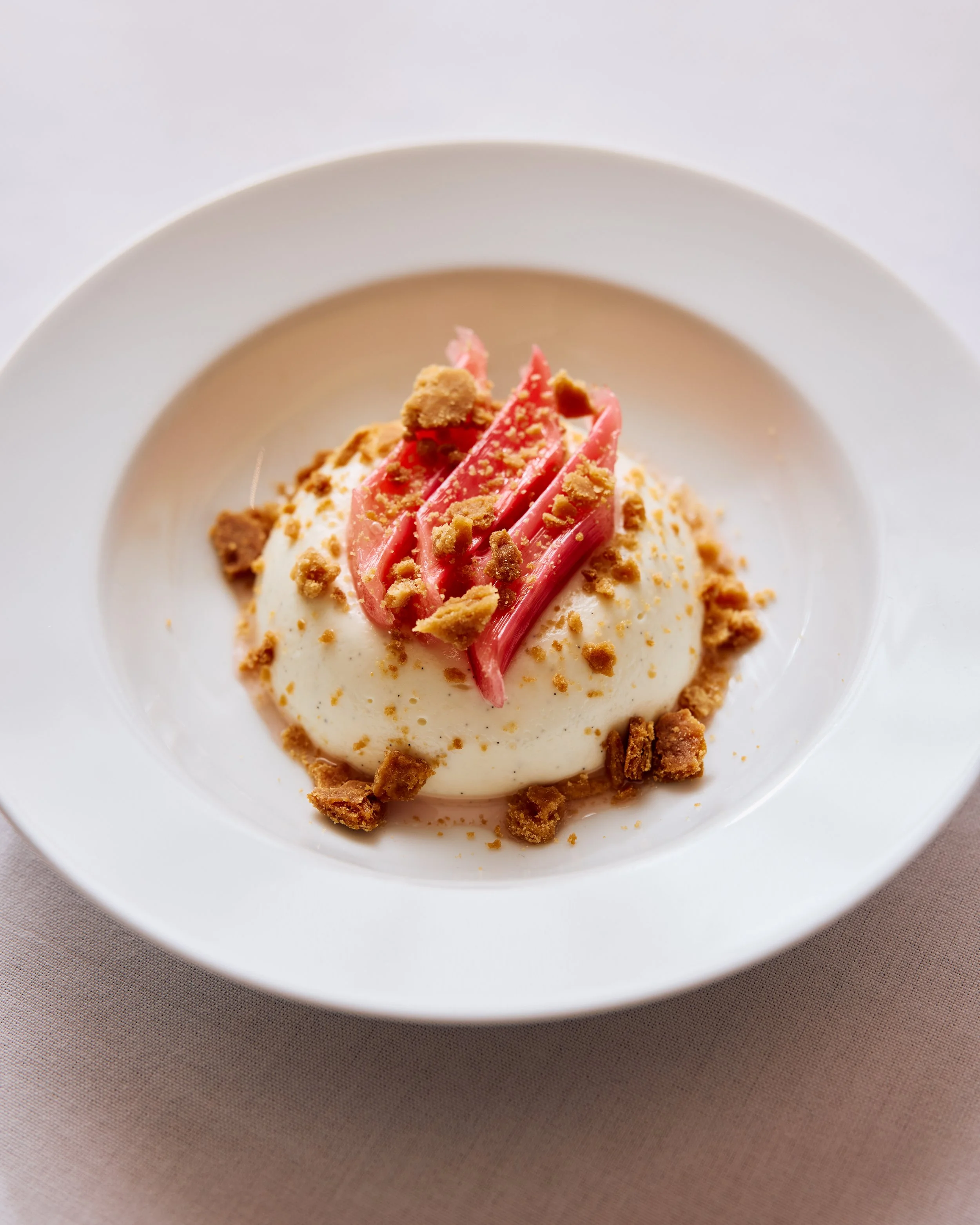 Panna cotta sits in a white bowl with bright pink sections of rhubarb and sprinkles of a brown cookie.