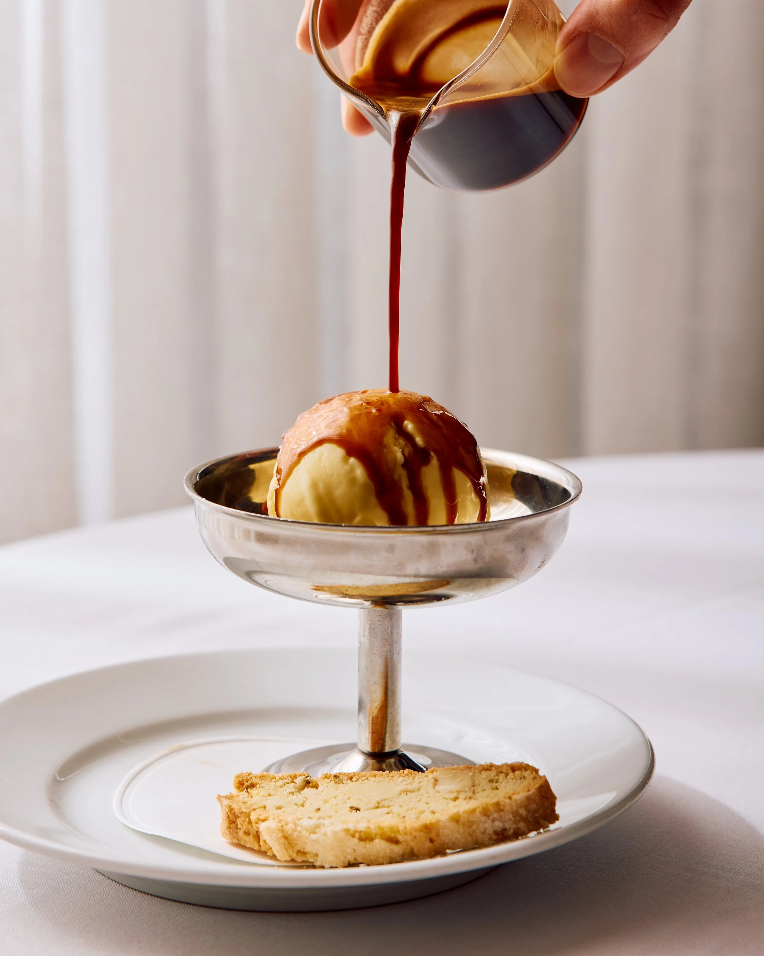 A silver coupe contains a scoop of fior di latte gelato. There is a hand pouring a shot of espresso over the gelato.