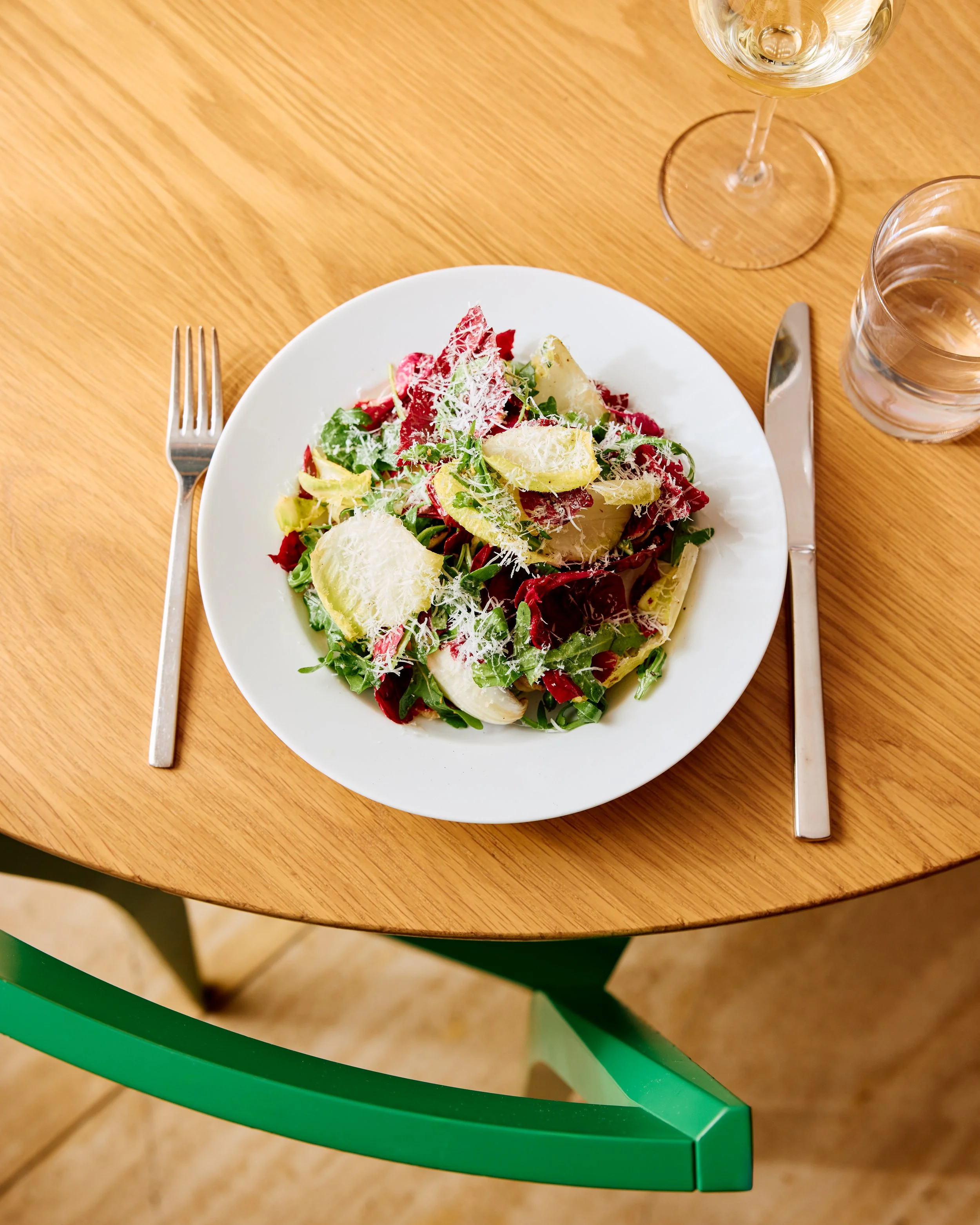 Image of a tricolore salad made of endive, arugula, radicchio, pecorino cheese in a red wine vinaigrette placed on a wooden table with a fork and knife flanking the sides.