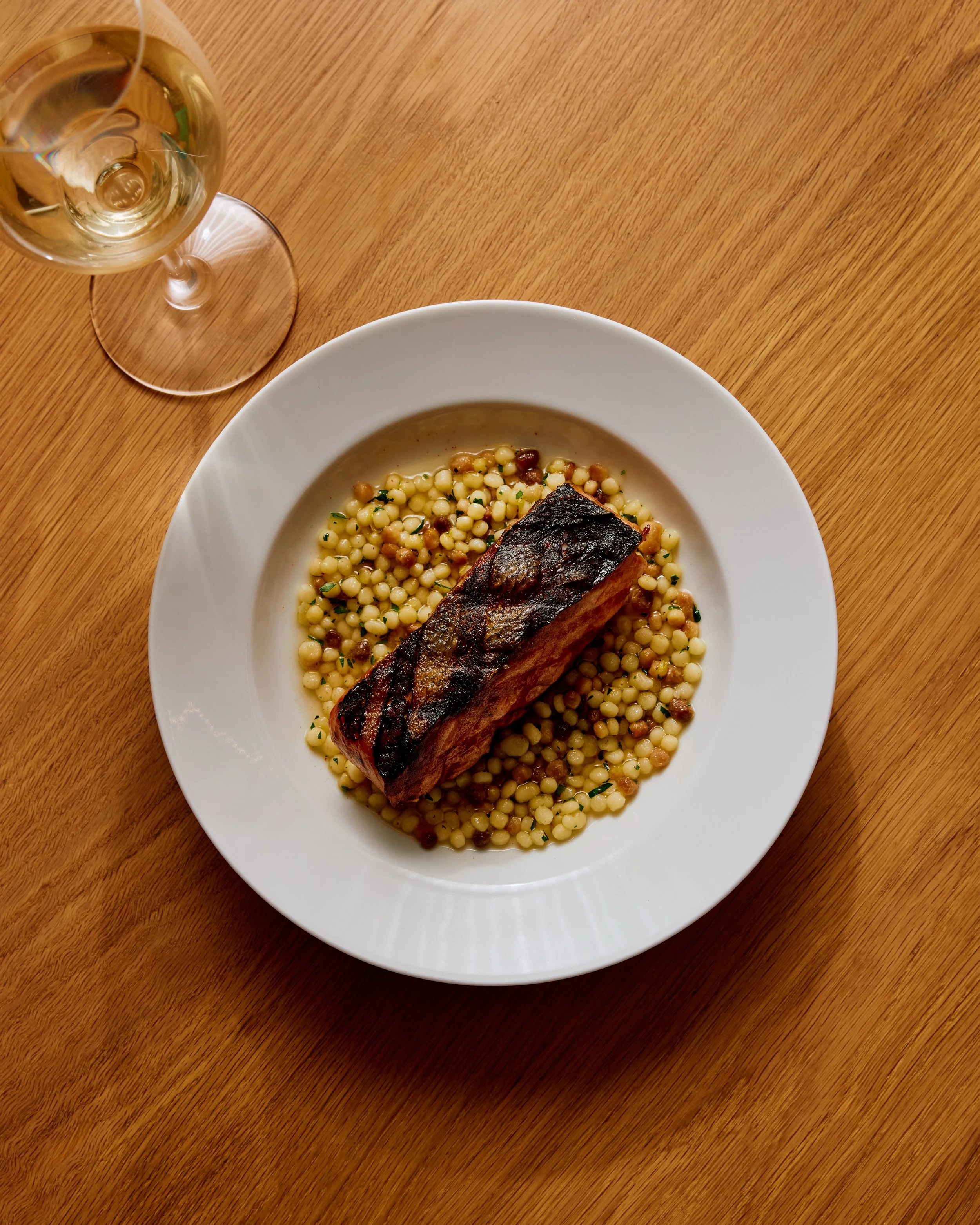 A bowl of grilled salmon sitting on top of a bed of small pearls of fregola on a wooden table with a glass of white wine placed on the left side.