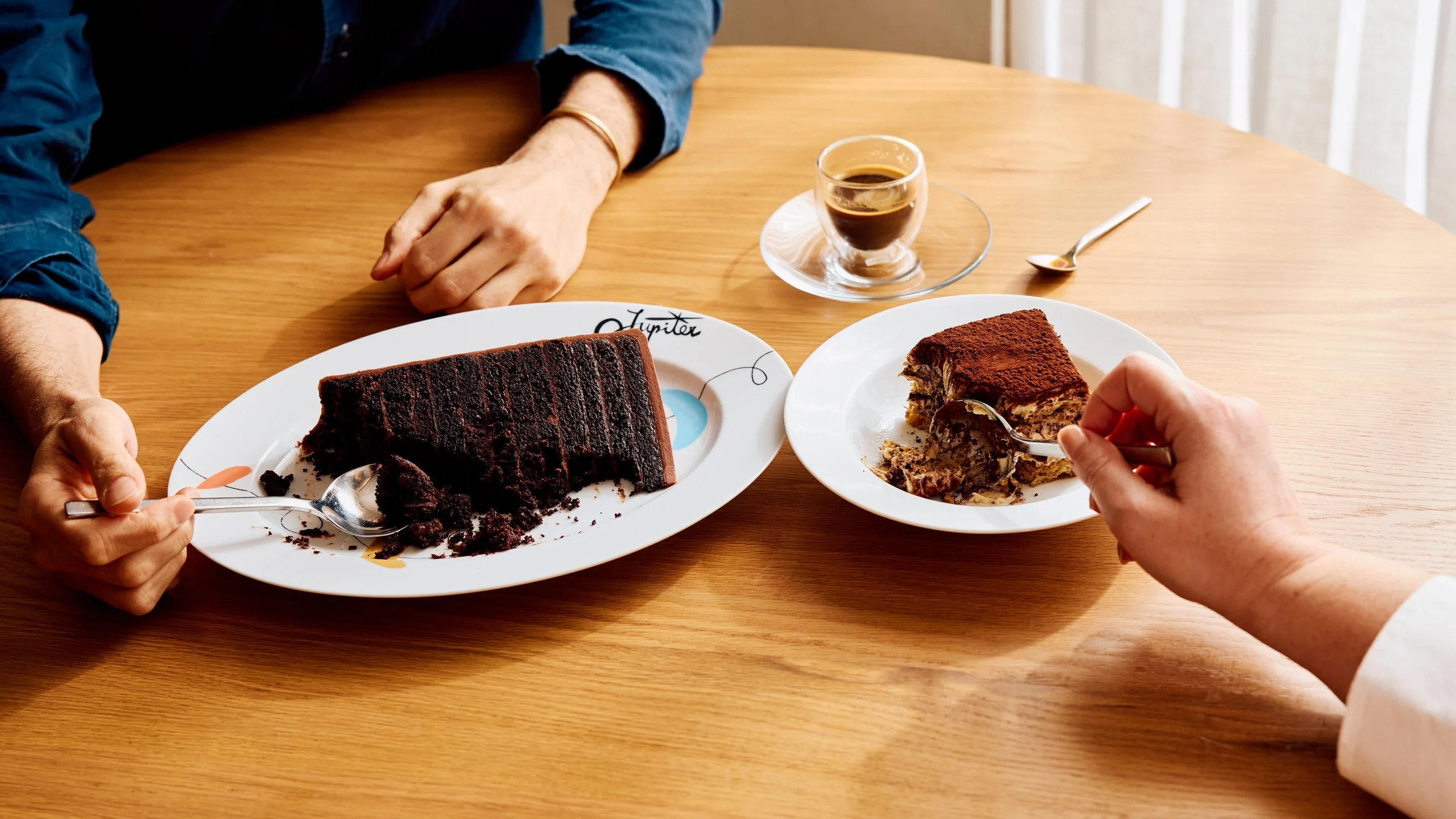 Two guests enjoy a table of desserts. One person is eating a chocolate cake made of 30 layers of cake and chocolate ganache. The other person enjoys a bowl of tiramisu. There is also a cup of espresso on the wooden oak table.