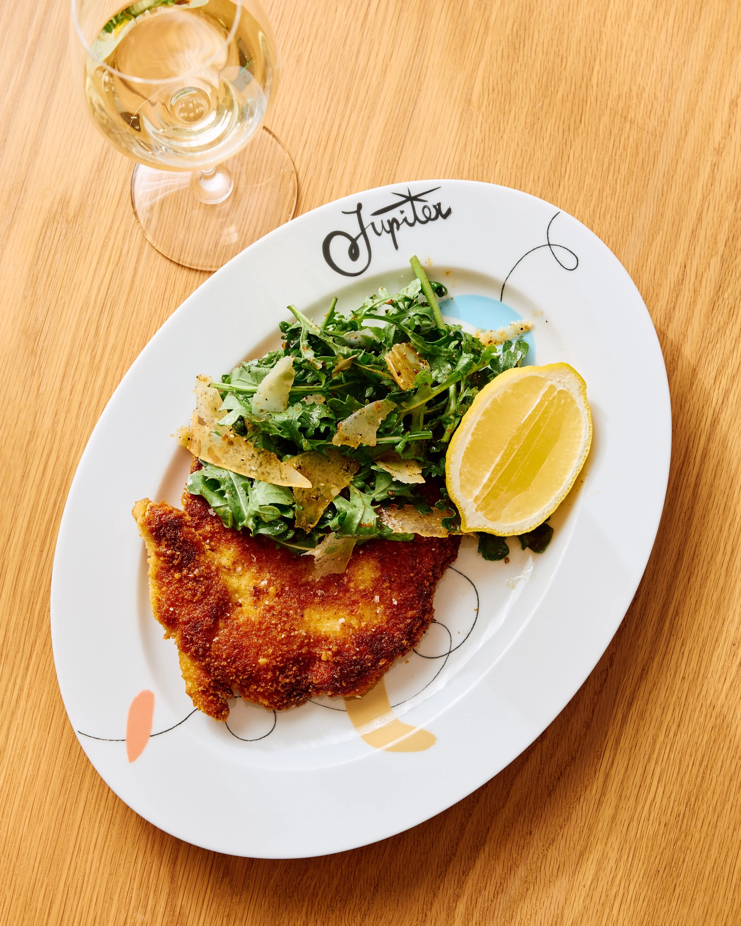 Crispy, lightly fried chicken sits beside arugula with aged balsamic and parmigiano regiano cheese sit on a while plate on top of a wooden table with a glass of white wine sitting next to it.