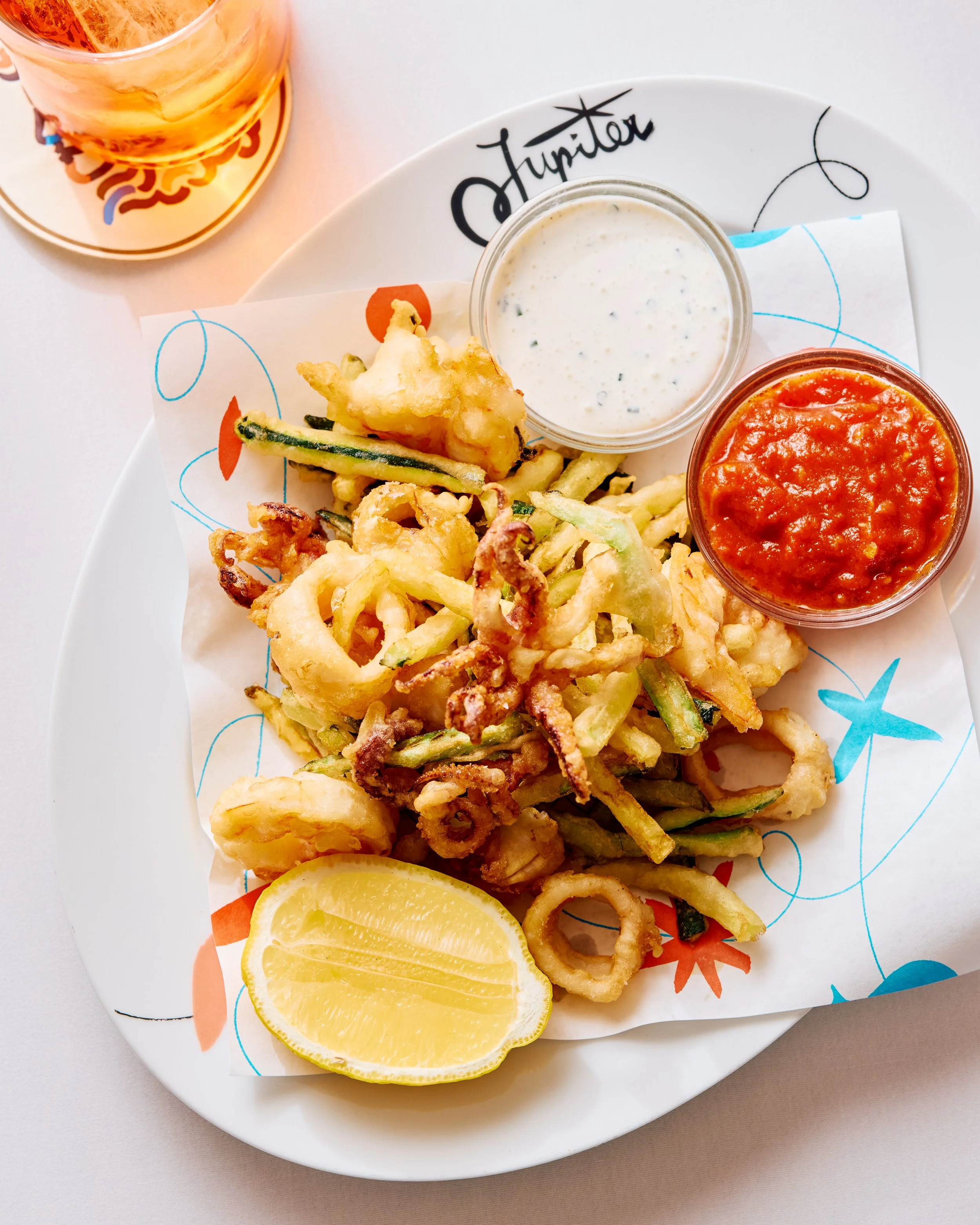 Golden fried calamari, shrimp and zucchini with a lemon wedge and a side of creme fraiche dressing, and pomodoro sauce. Sitting next to the plate is an aperol spritz.