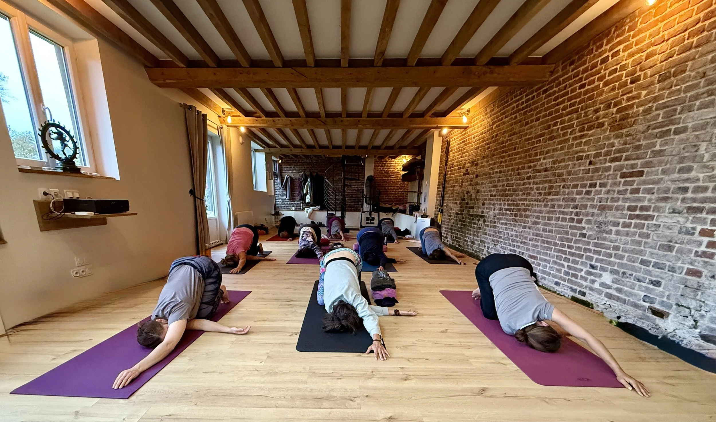 Cours de yoga tous niveaux en Normandie, groupe de pratiquants en posture de relaxation dans une salle lumineuse aux murs en briques et parquet en bois.