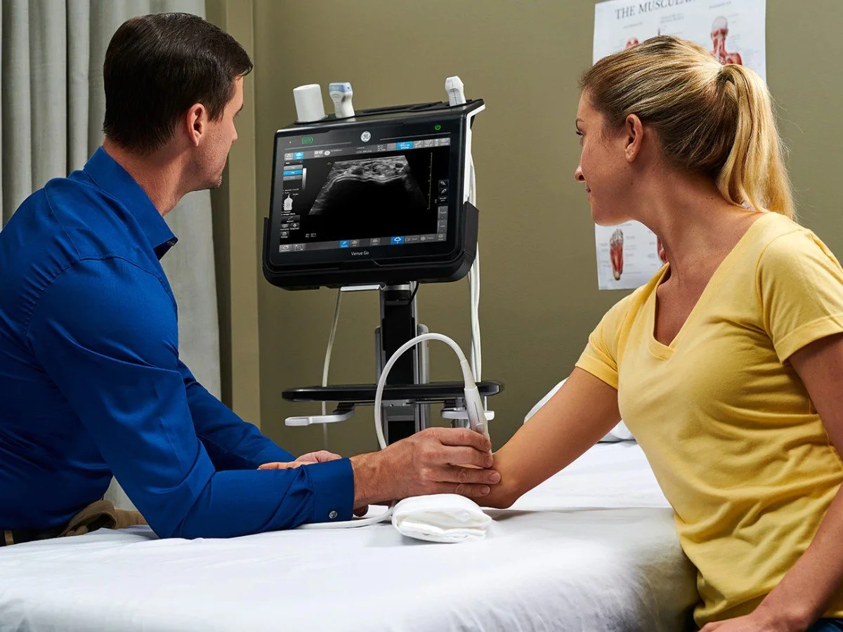 A medical professional using an ultrasound machine on a patient's arm in a clinic setting.