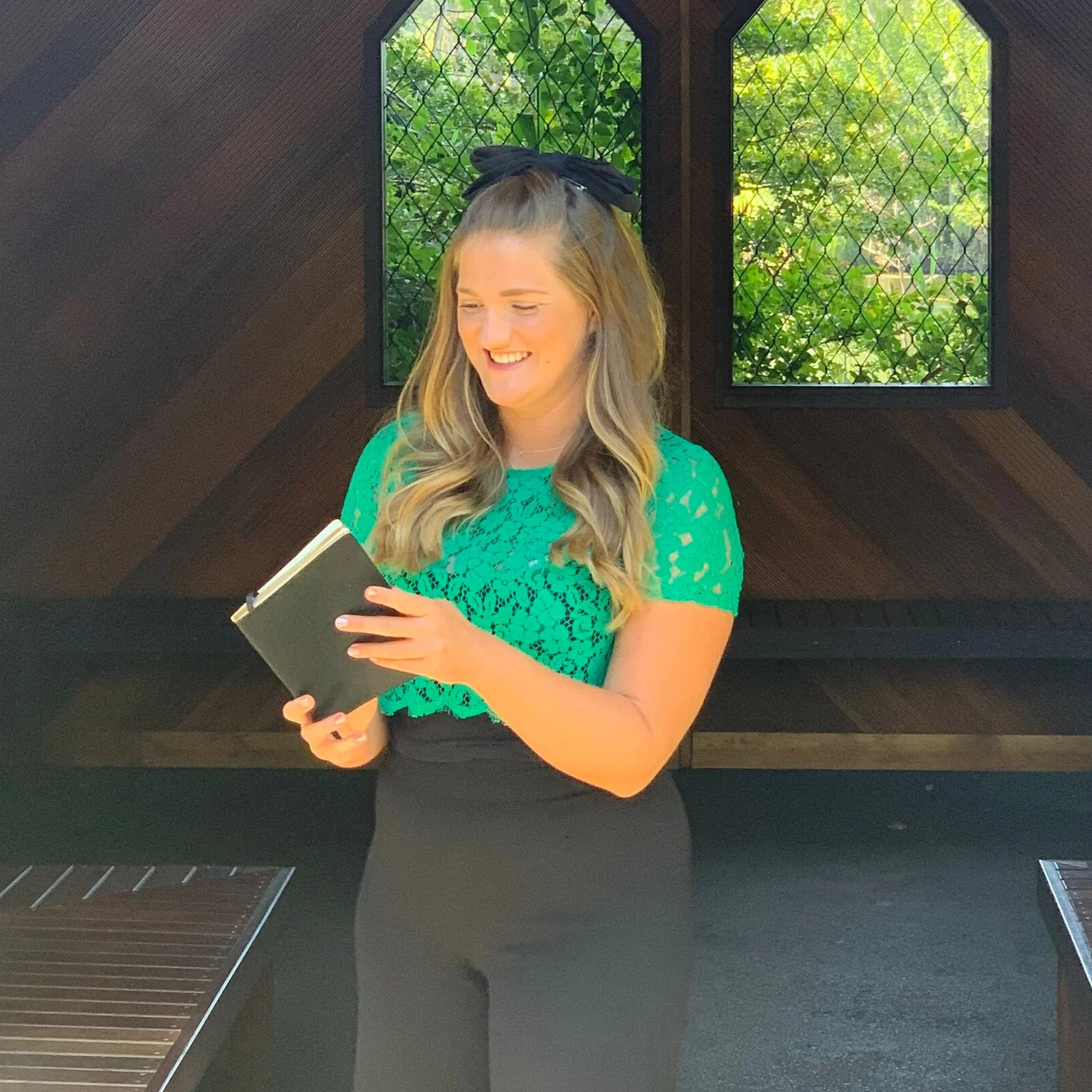 A young woman with long blonde hair, wearing a black headband and a bright green lace top, is smiling and reading a black notebook. She is standing indoors with dark wooden walls and two windows with green foliage outside.