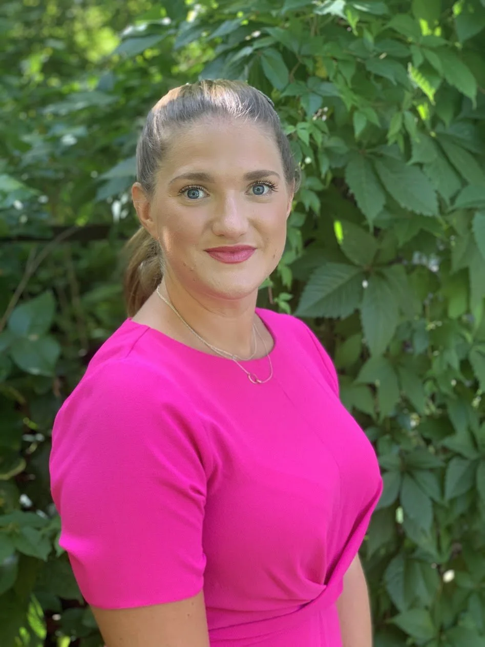 A woman with light skin, blue eyes, and brown hair tied back in a ponytail, wearing a bright pink dress and a delicate necklace, standing outdoors in front of green leafy foliage.