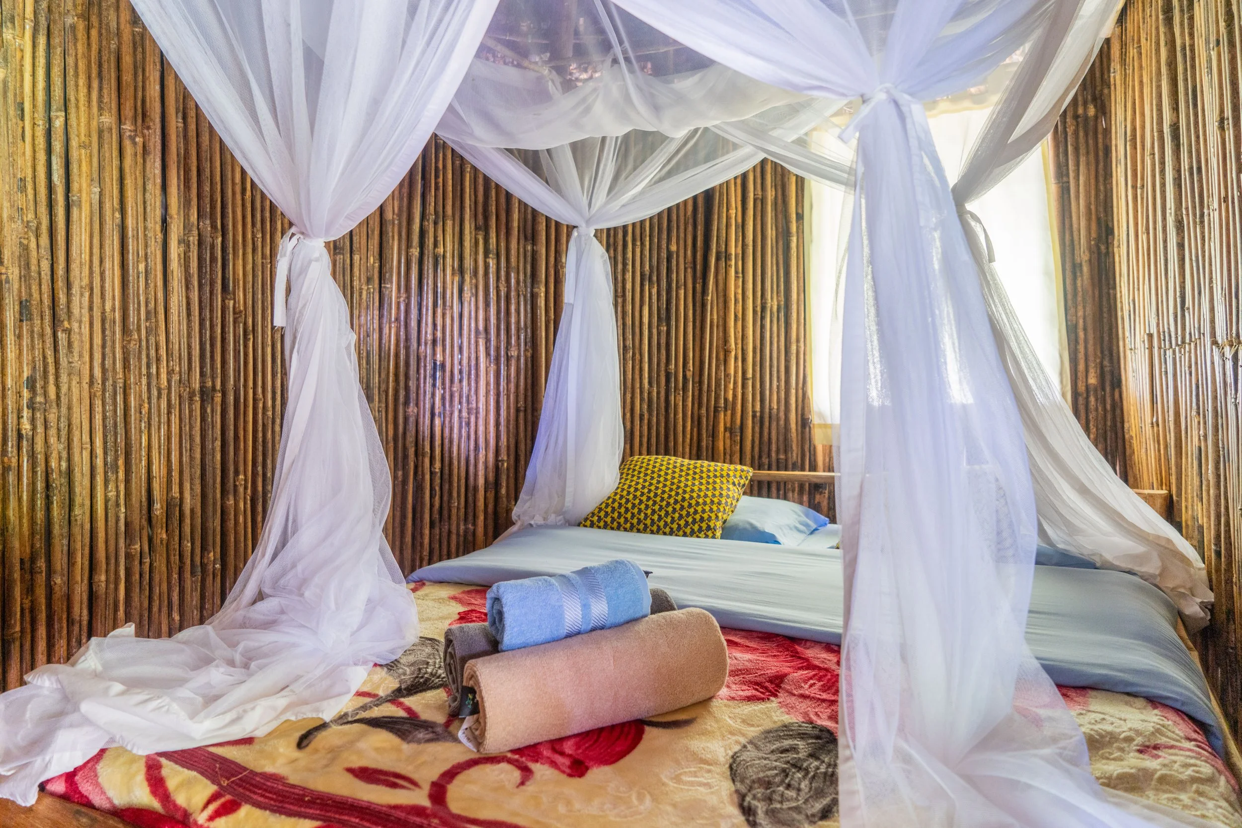 A bedroom with a canopy bed with white fabric draped around the wooden frame, a bed with a floral blanket, pillows, and rolled towels, and sunlight coming through the window.