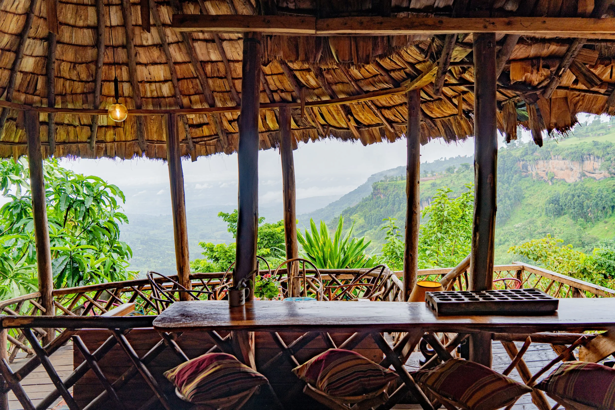 A rustic open-air wooden deck with a thatched roof overlooking green hills and mountains.
