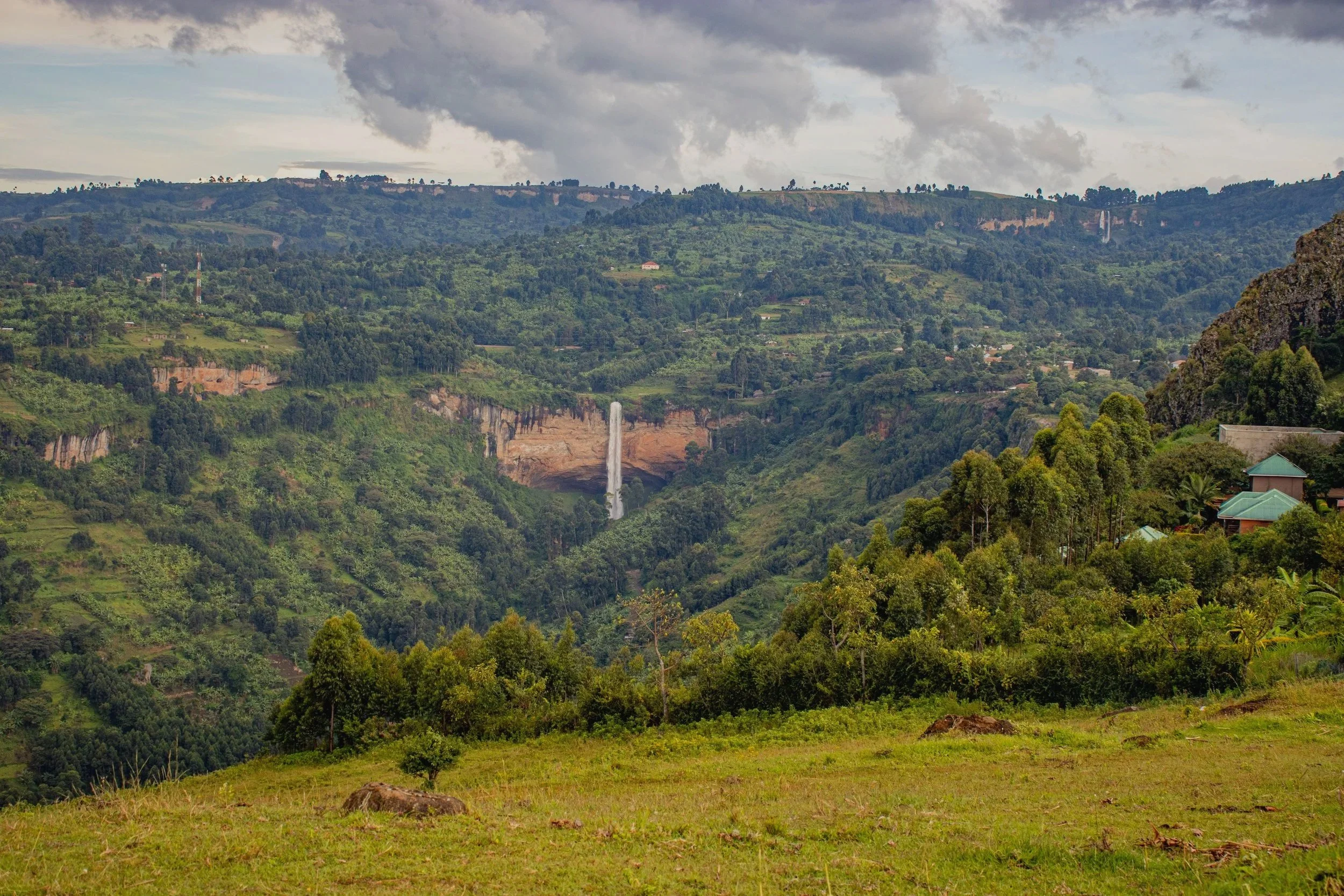 Scenic view of lush green hills with a waterfall in the distance, under a cloudy sky.