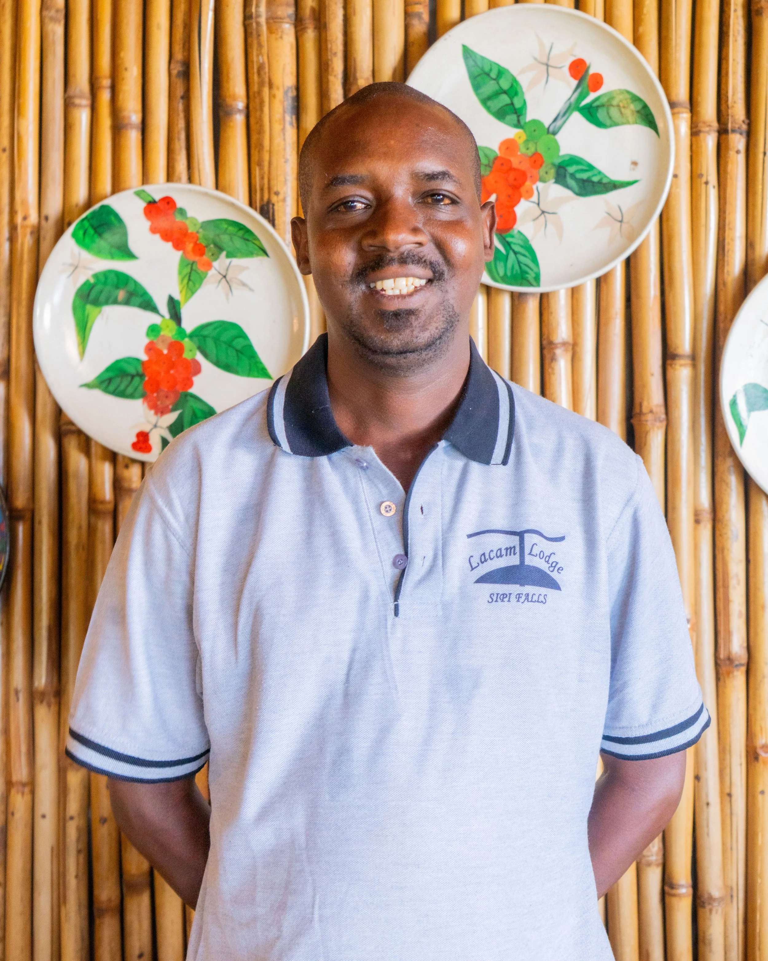A man standing in front of a bamboo wall, smiling, wearing a gray polo shirt with "Lacam Lodge Sipili Falls" embroidered on it, with decorative plates featuring green leaves and red berries hanging behind him.