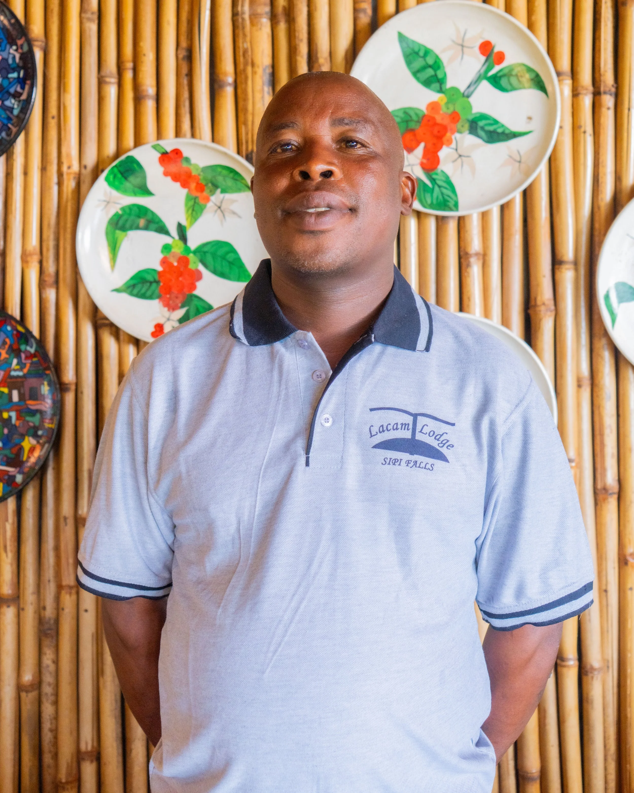 A man standing in front of a bamboo wall decorated with painted plates featuring green leaves and red berries, wearing a gray polo shirt with a logo on the chest that says Lacam Lodge, Silver Falls.