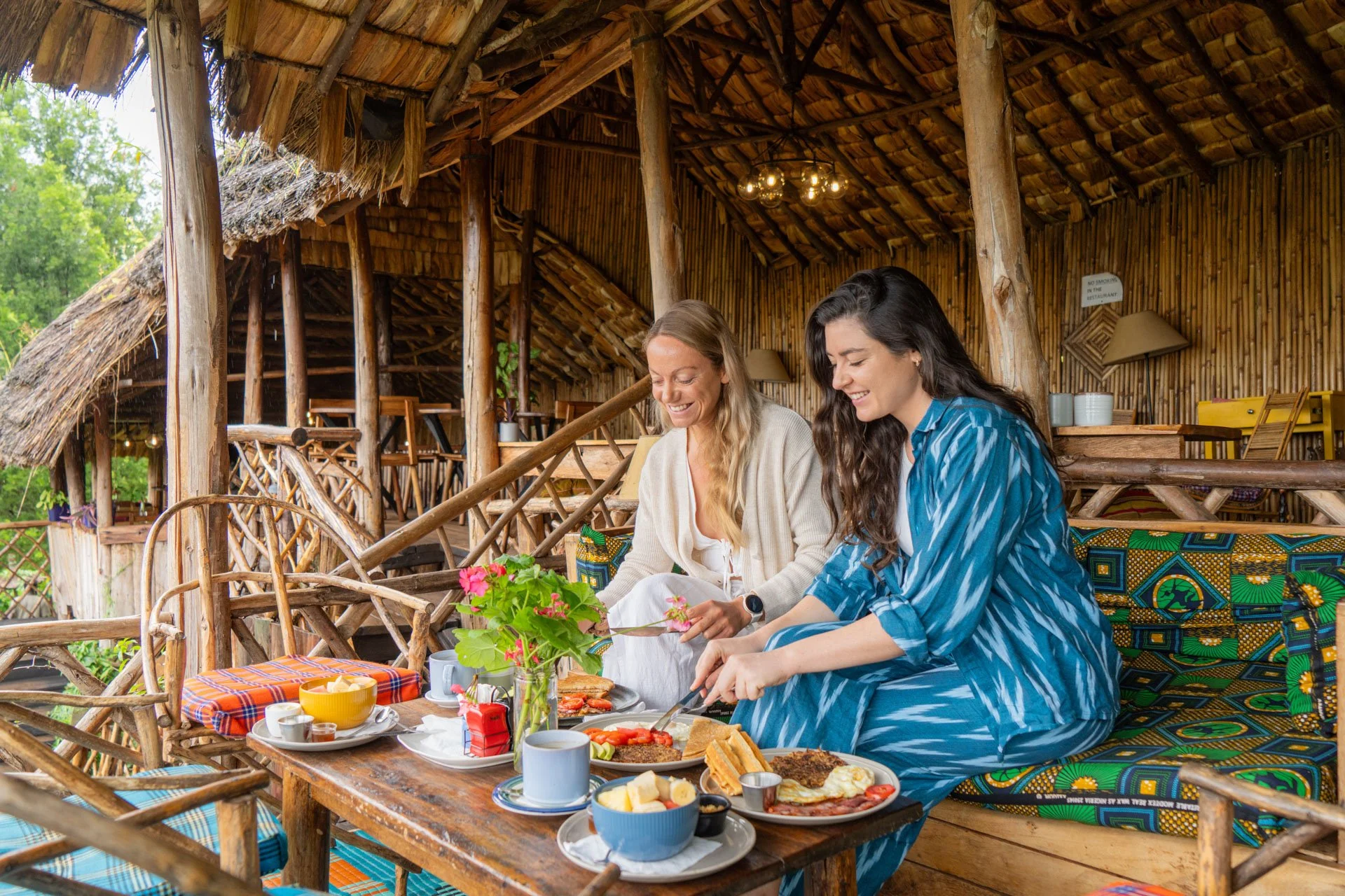 Two women sitting at a wooden table enjoying breakfast in a rustic outdoor setting with bamboo walls and thatched roof, smiling and cutting food together.