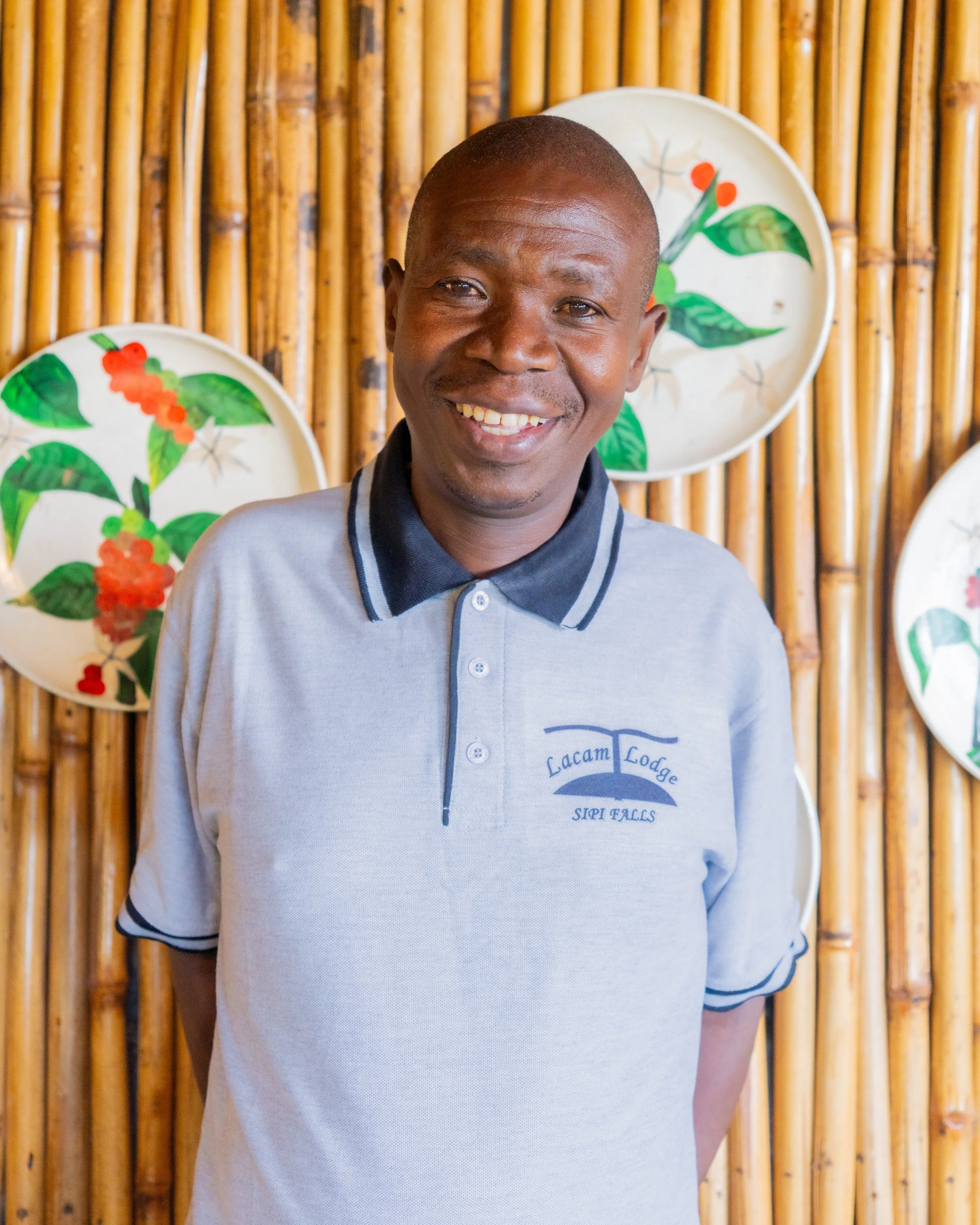 A smiling man in a gray polo shirt standing in front of a bamboo wall with decorative plates featuring painted green leaves and orange berries.