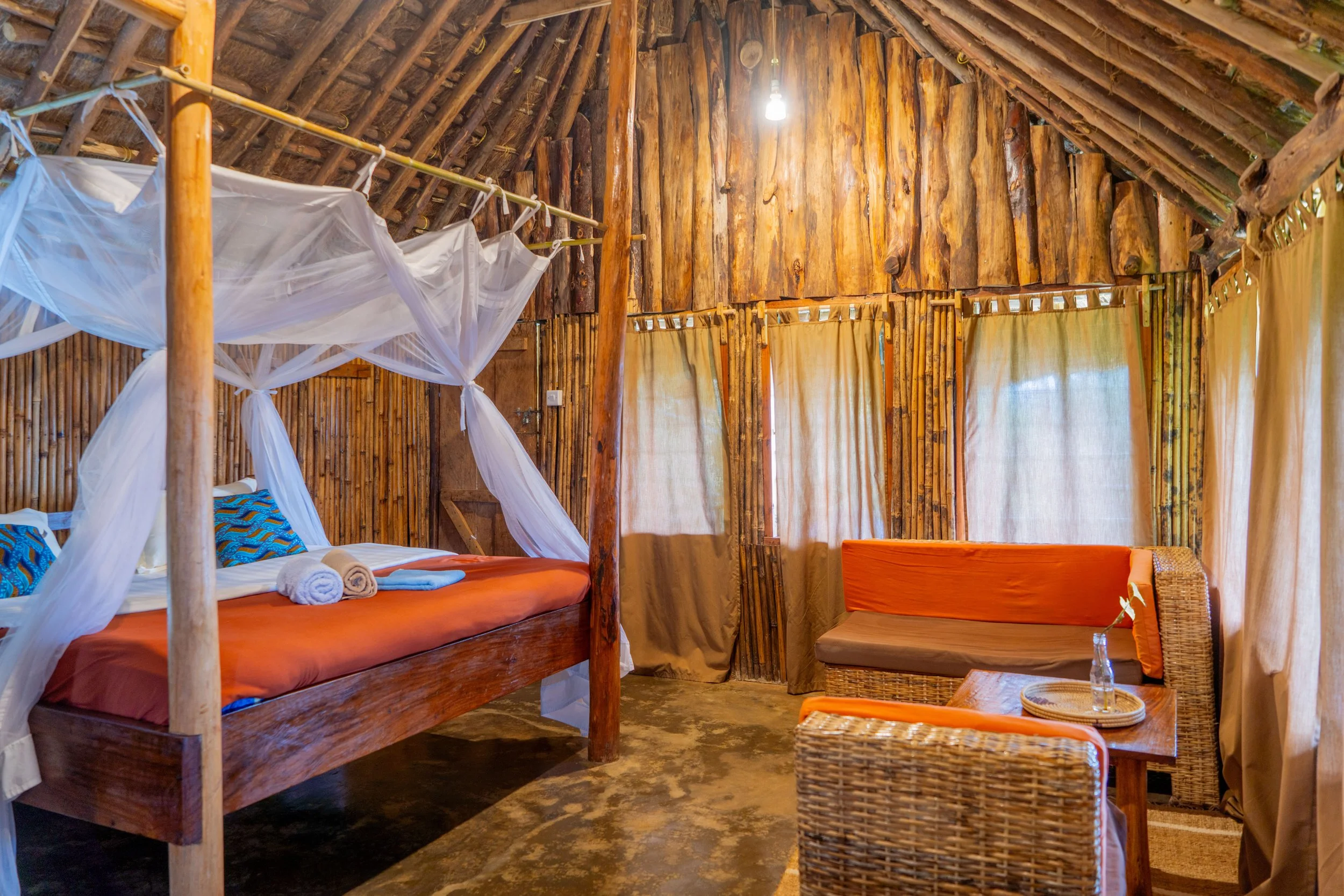 A rustic bedroom with wooden walls and ceiling, featuring a four-poster bed with white mosquito netting, orange bedding, and rolled towels on top. There is a sitting area with wicker furniture and a small wooden table with a glass bottle and tray, with windows covered by beige curtains.