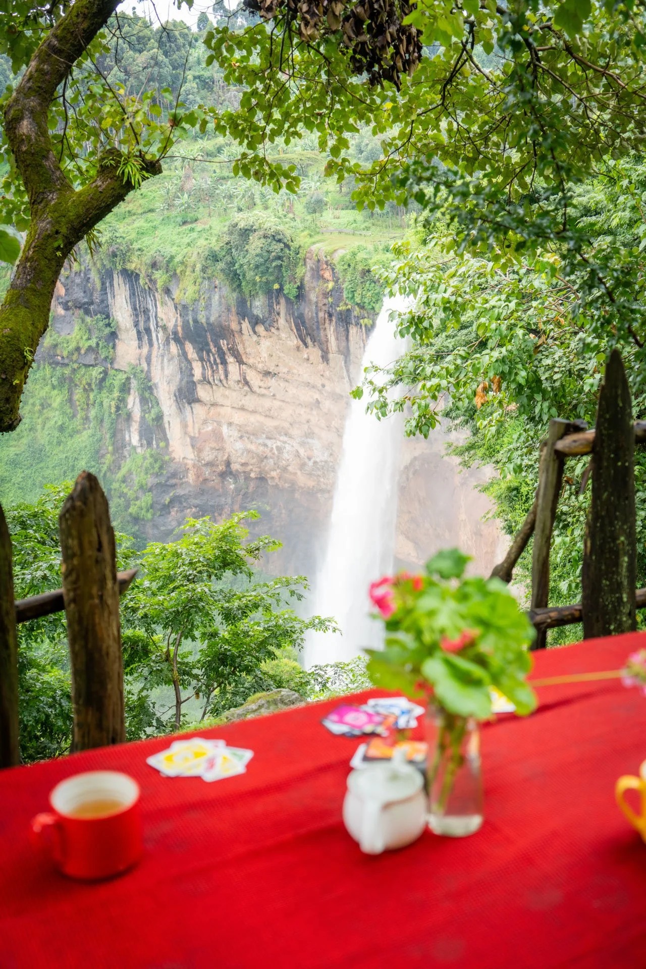 A view of a waterfall through a lush green forest, seen from a table with a red tablecloth, a small flower vase, and some papers or cards.