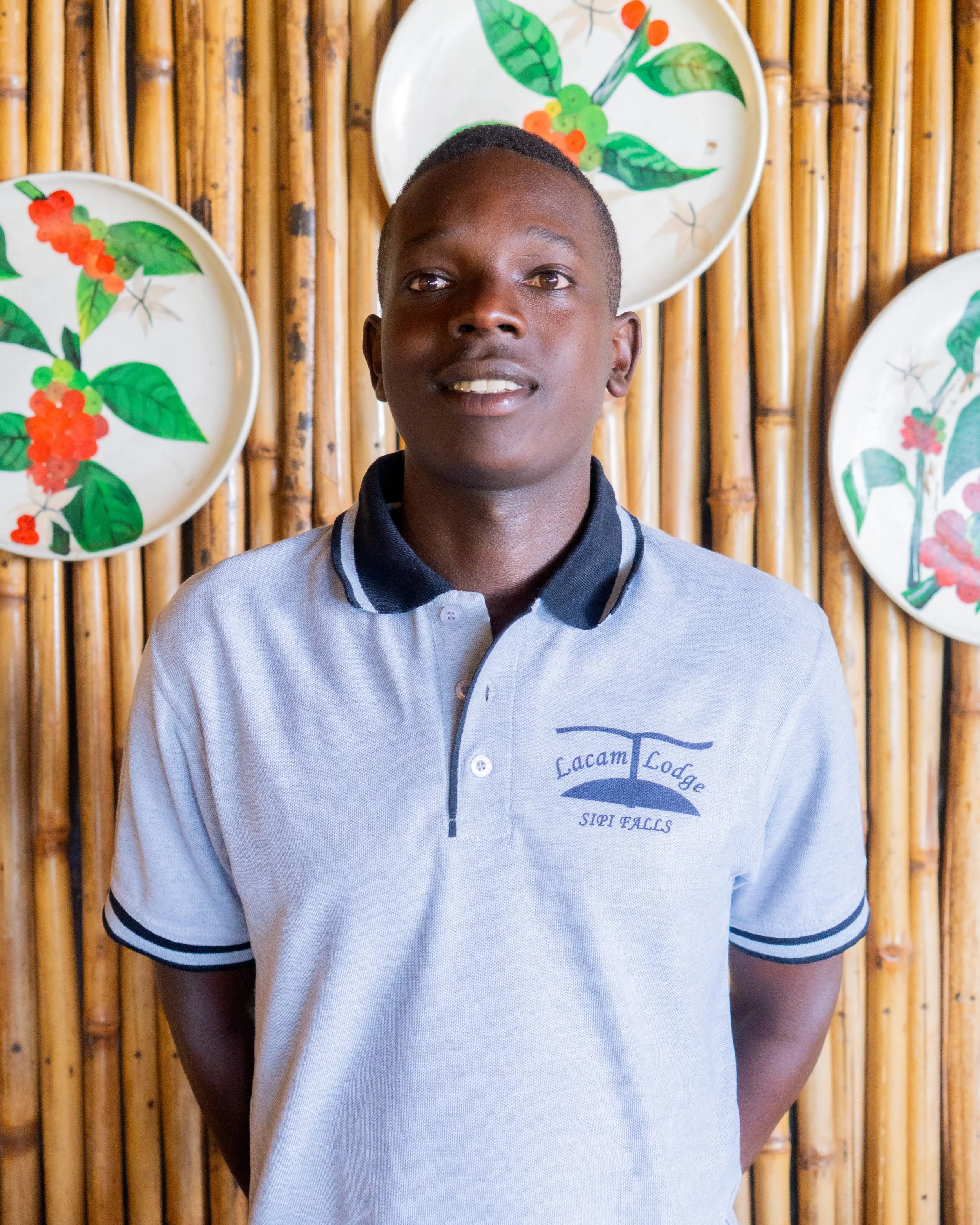 A young man with dark skin and short hair, wearing a light gray polo shirt with a logo, standing in front of a bamboo wall decorated with four painted plates featuring green leaves and red berries.