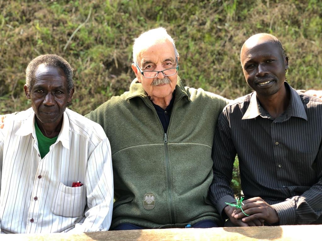 Three men sitting outdoors in front of a green, hilly background, with one elderly man at the center, wearing glasses and a green zip-up jacket, flanked by two younger men, all looking at the camera.