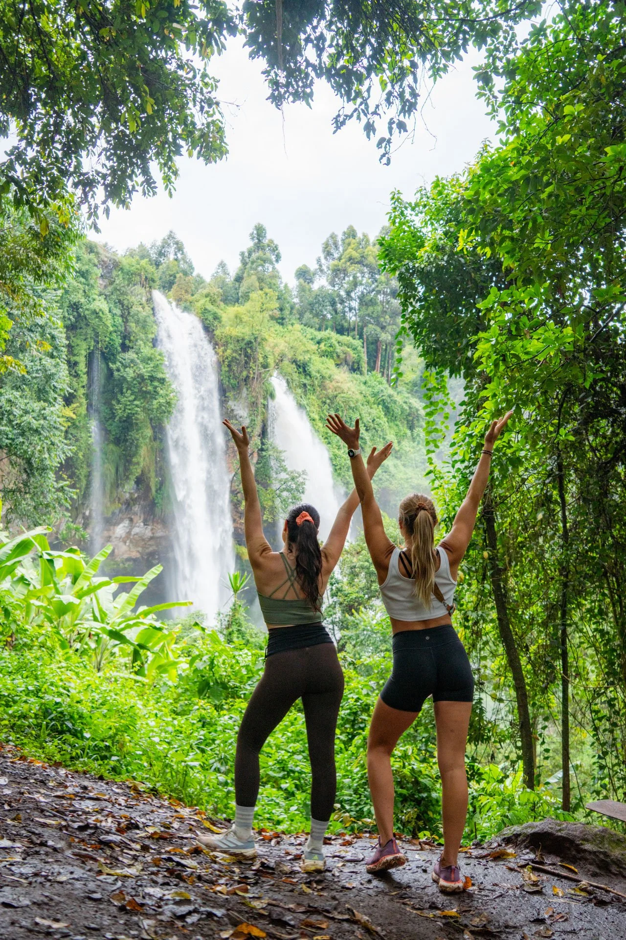 Two women with arms raised stand in a lush green forest near a waterfall.