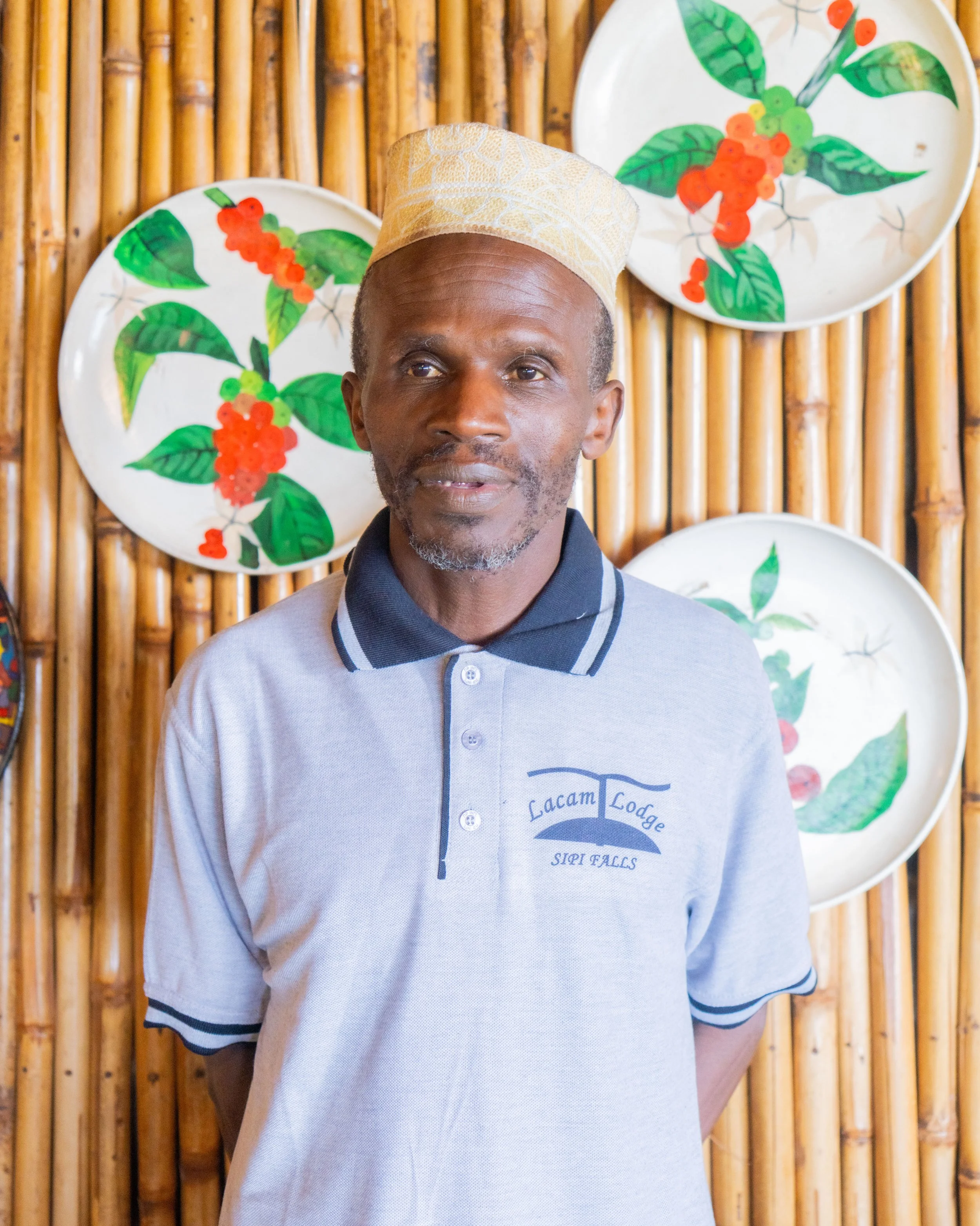 A man with a beard wearing a gray Lacam Lodge polo shirt and a traditional beige hat standing in front of a bamboo wall decorated with white plates featuring colorful green leaves and red berries.
