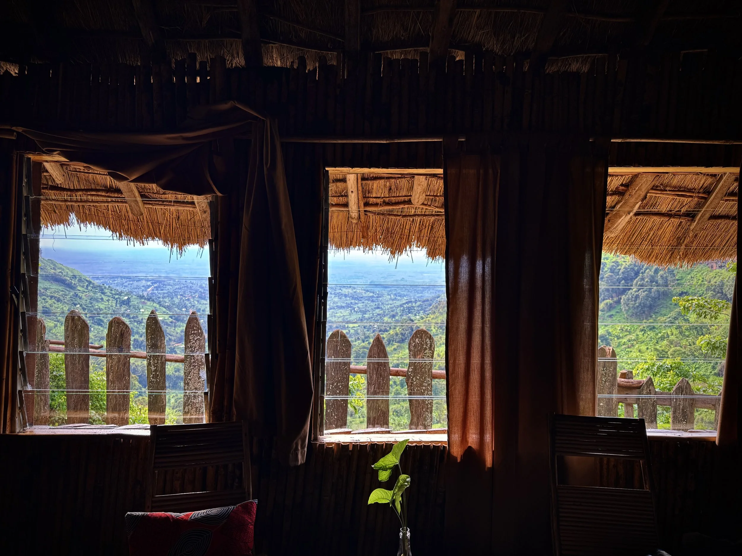 Interior of a rustic wooden hut with open windows showing a lush green landscape and mountains in the distance.