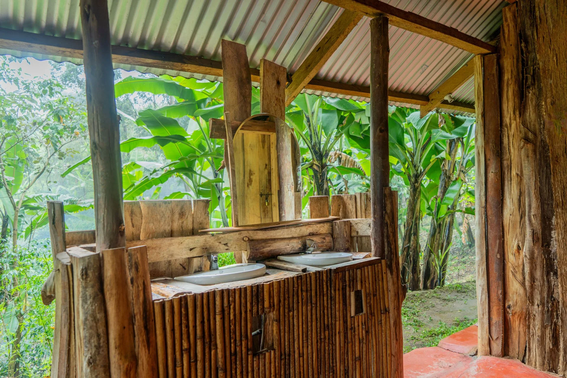 Wooden outdoor bathroom with two sinks, mirror, and lush green banana trees outside.