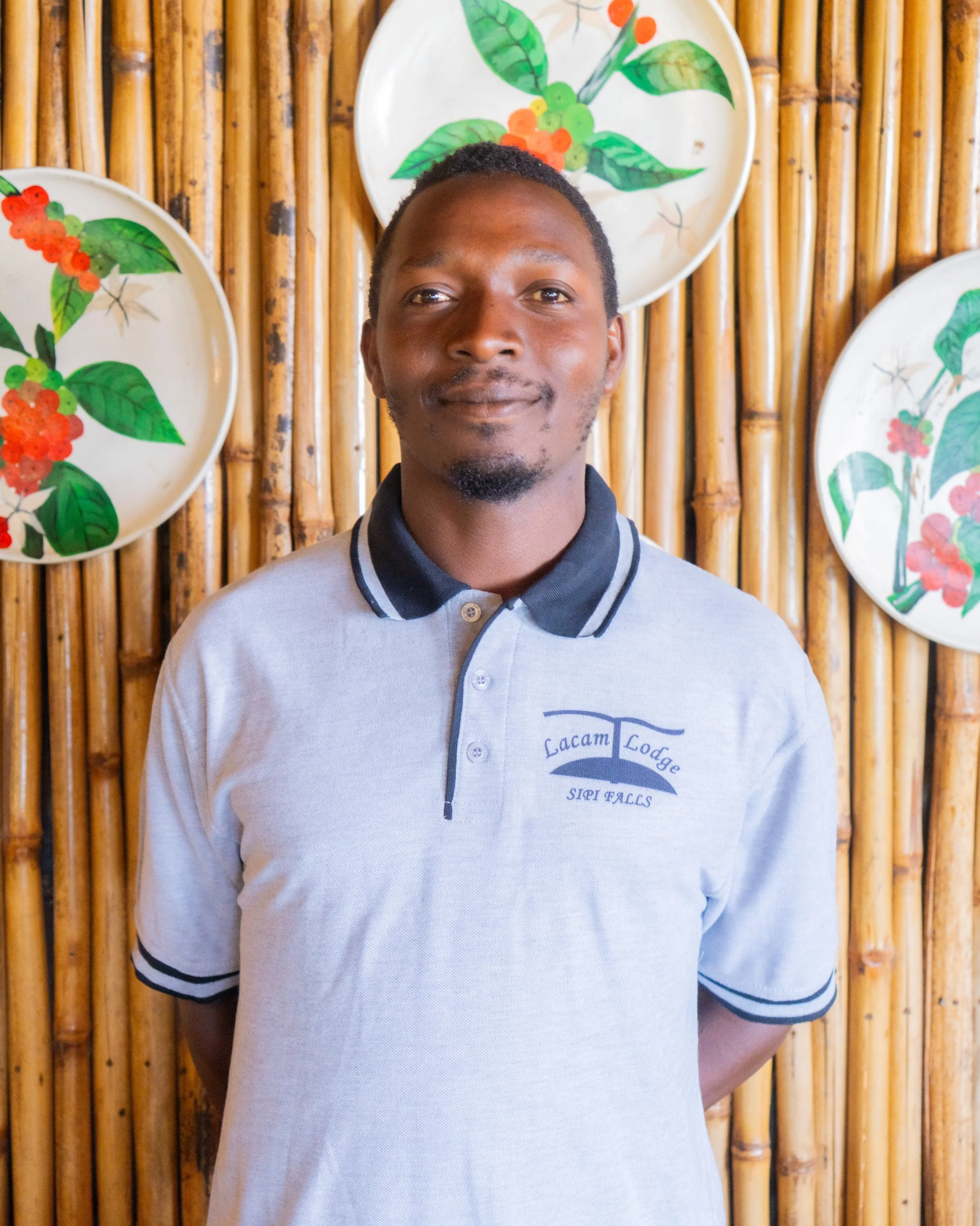 A man standing in front of a bamboo wall decorated with painted ceramic plates featuring green leaves and red berries. He is wearing a gray polo shirt with dark collar and sleeve trim, with greeting text on it.