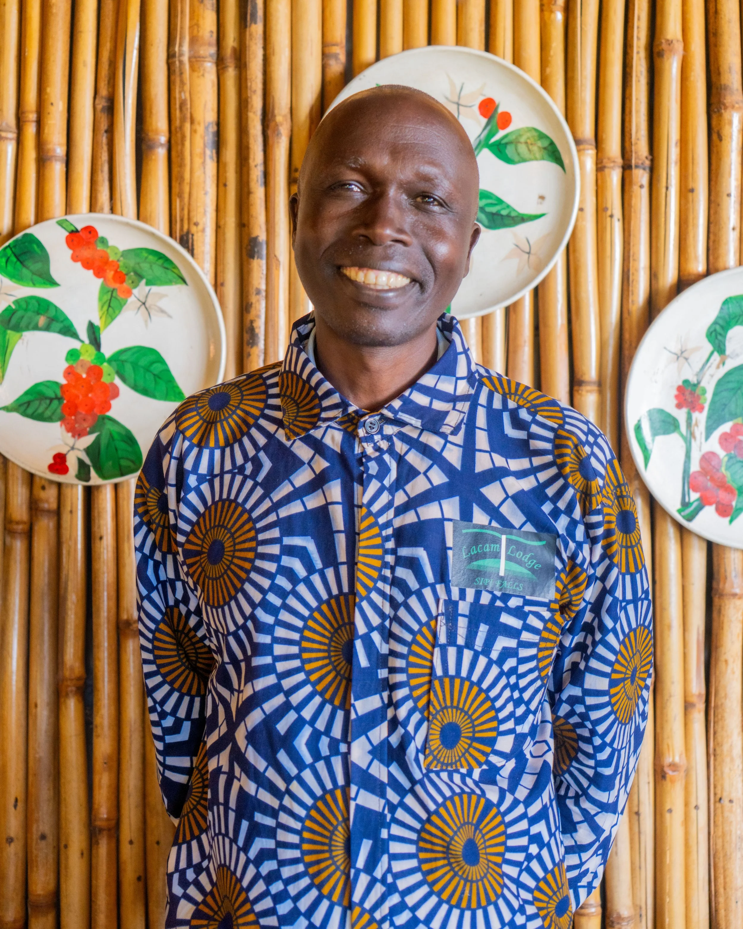 Smiling man in colorful patterned shirt standing in front of bamboo wall with decorative plates with green leaves and red berries.
