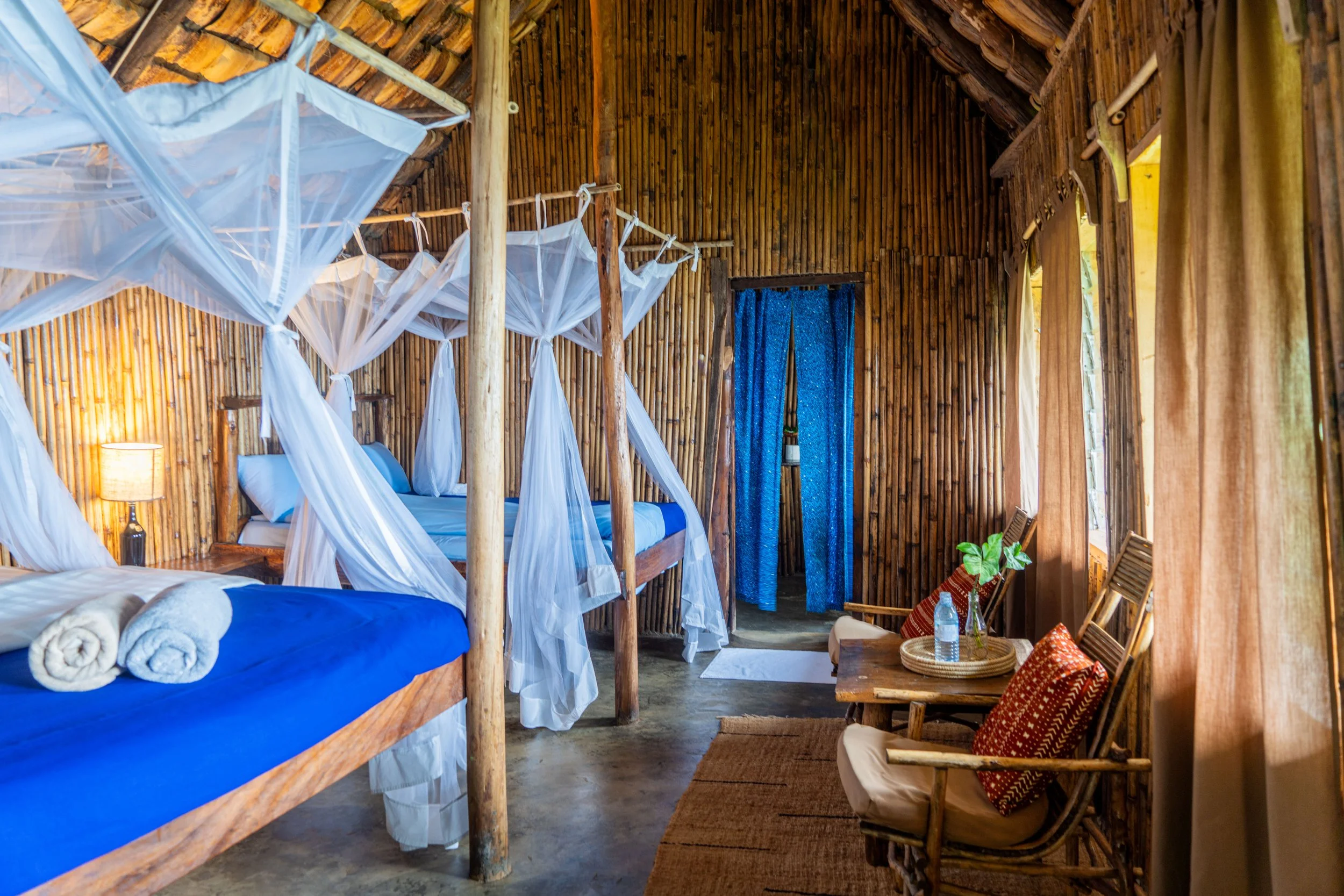 Interior of a rustic wooden cabin bedroom with two beds, one with blue bedding and two rolled towels, surrounded by mosquito nets, a small wooden table with two chairs, and a doorway with blue curtains.