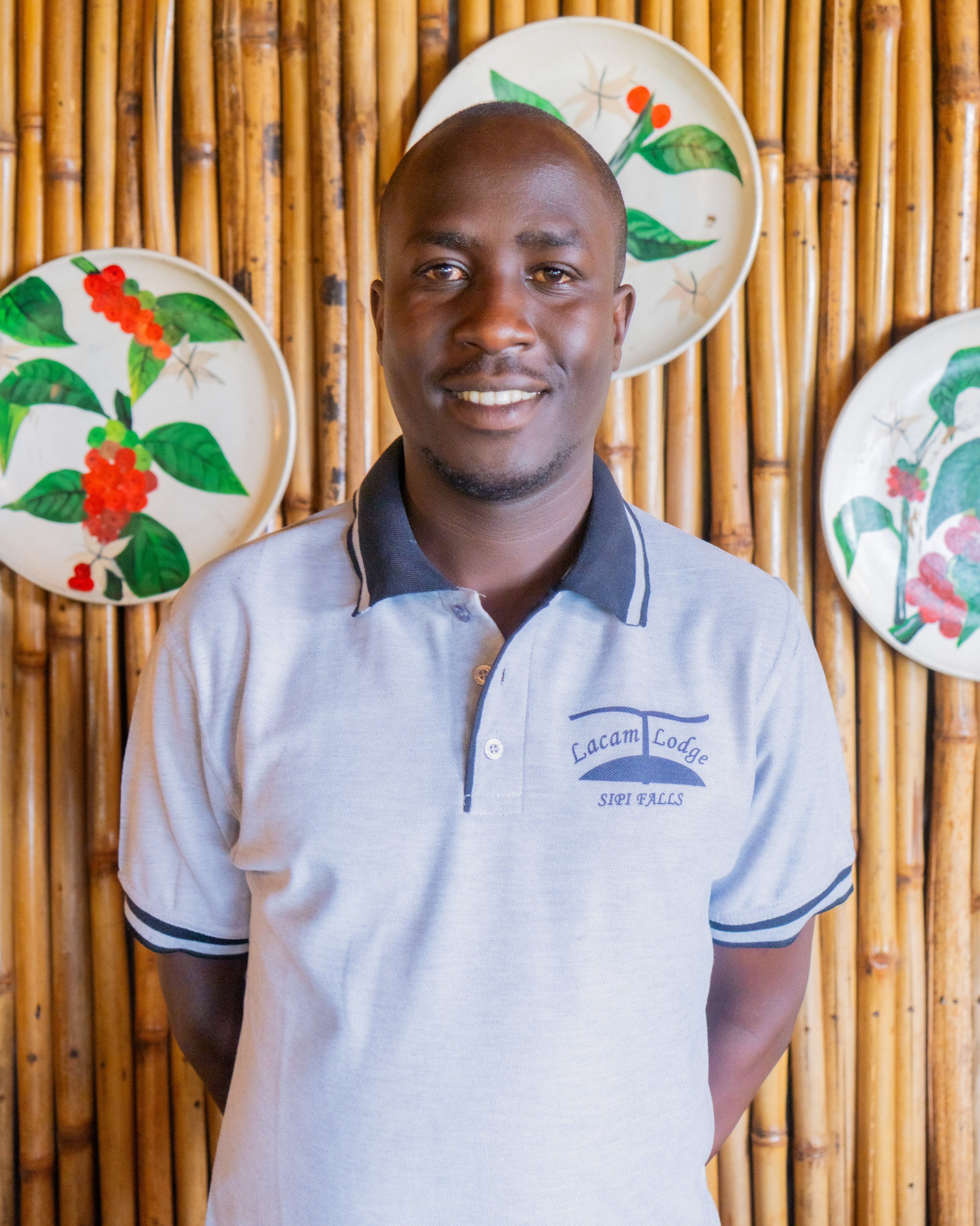 A man standing in front of a bamboo wall decorated with white plates featuring colorful botanical designs, wearing a light gray polo shirt with the embroidered logo of Lacam Lodge, Sip Fall.