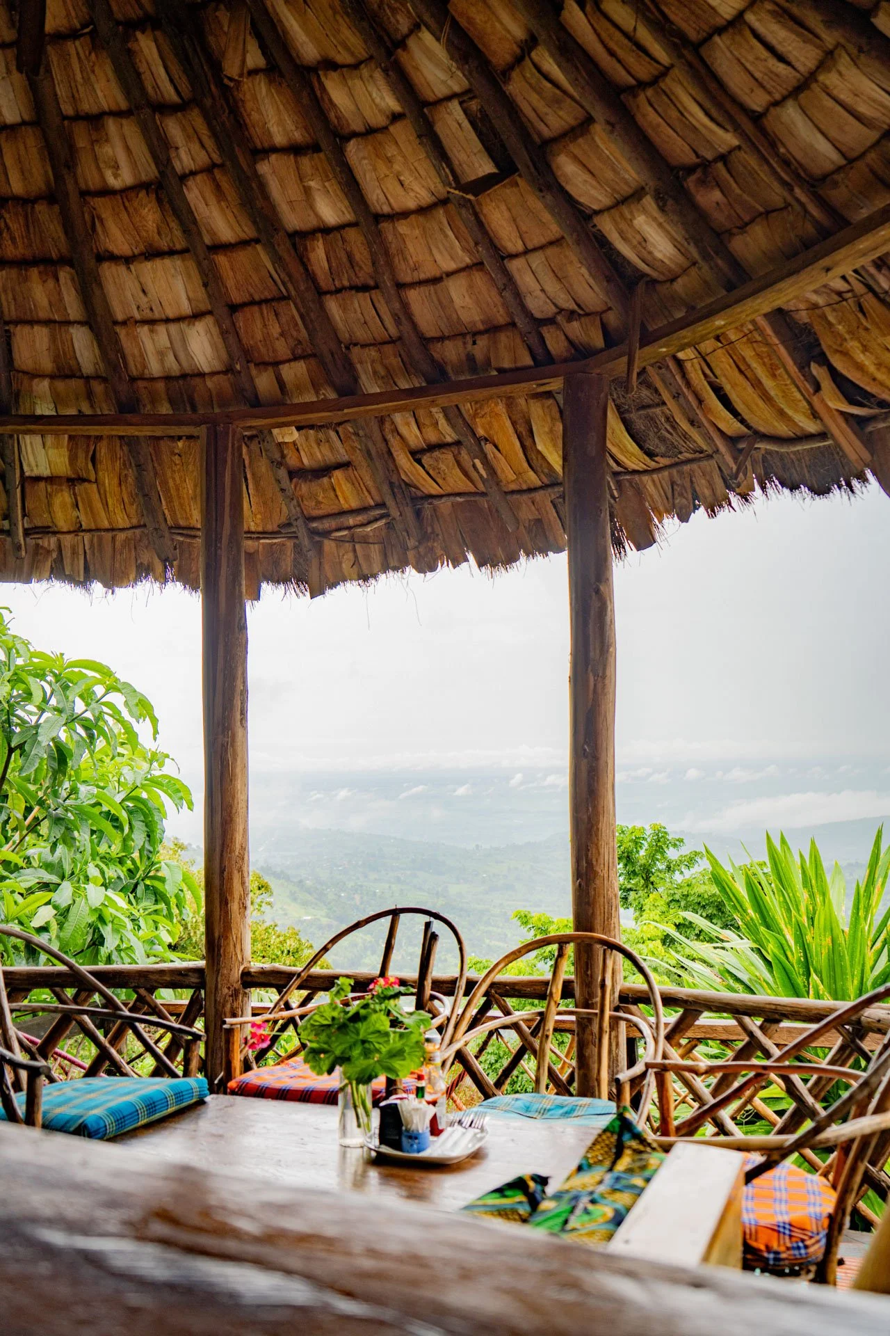 A rustic outdoor dining area with a wooden table and chairs, under a thatched roof, overlooking lush greenery and a distant mountain landscape.